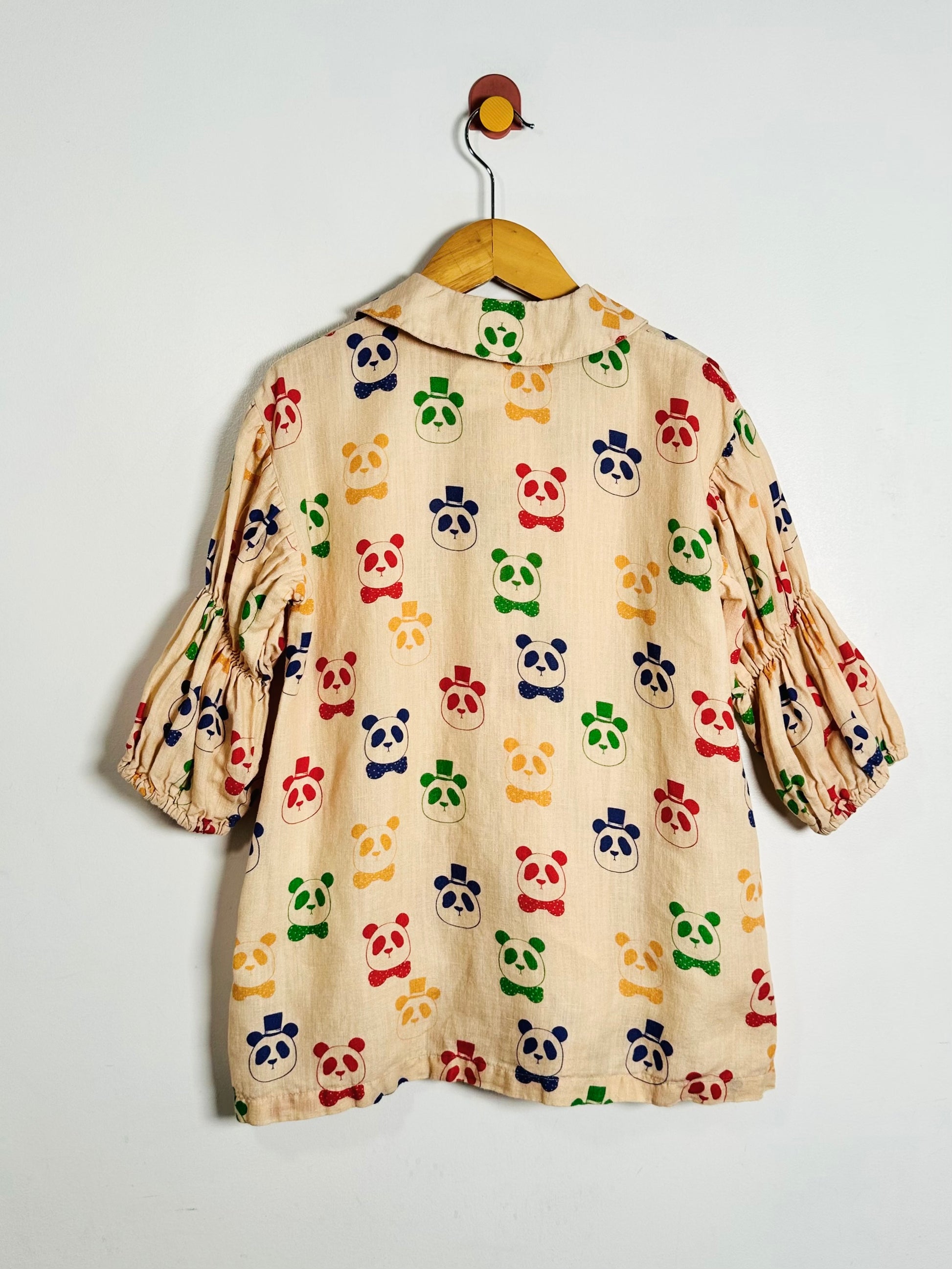 A beige cotton dress with a colorful panda print, hanging on a wooden hanger against a plain white wall.