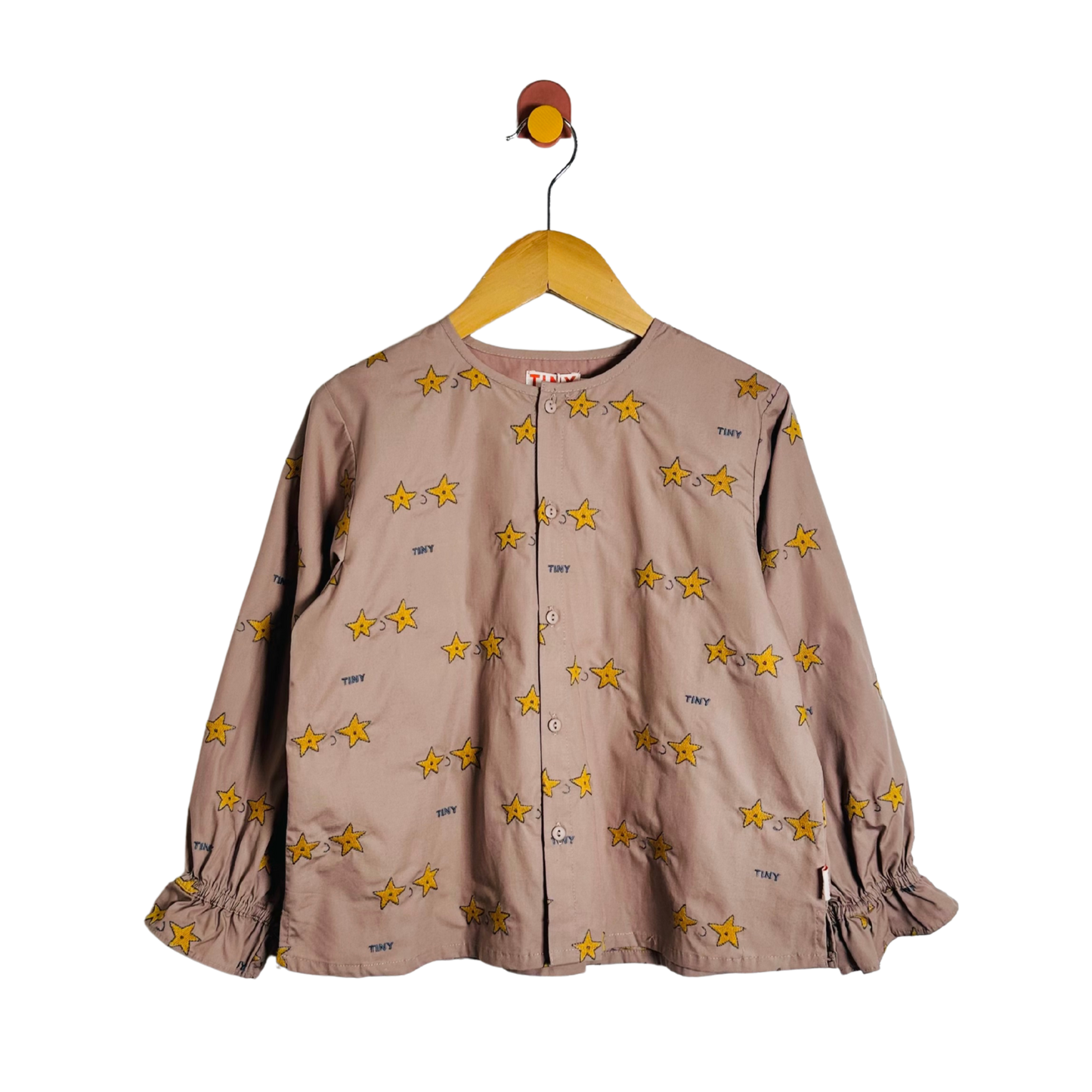 A children's long-sleeve button-front top in a muted taupe-brown color, decorated with an all-over print of yellow stars outlined in black. The top features elasticized cuffs and is shown hanging on a wooden hanger against a plain white background.