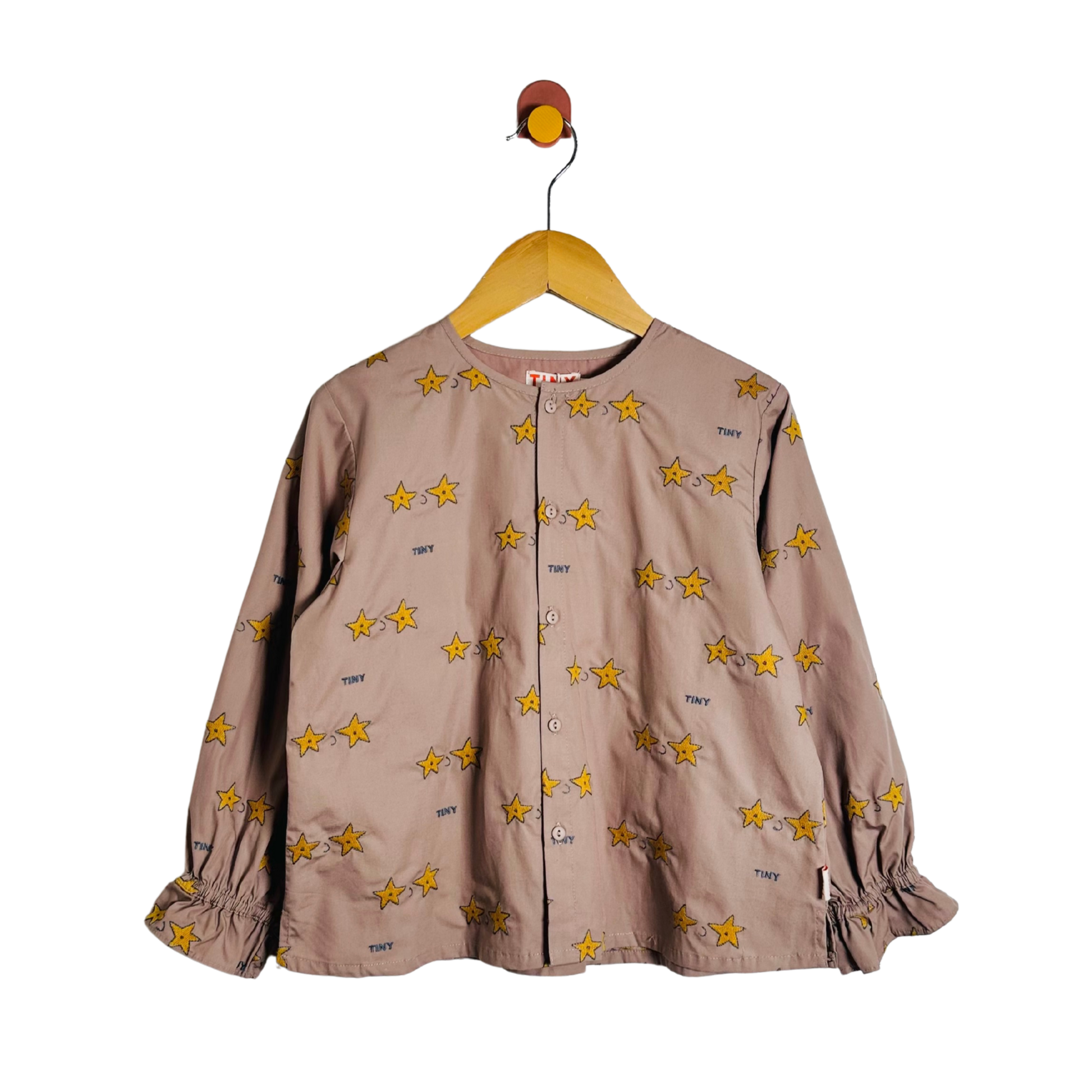 A children's long-sleeve button-front top in a muted taupe-brown color, decorated with an all-over print of yellow stars outlined in black. The top features elasticized cuffs and is shown hanging on a wooden hanger against a plain white background.