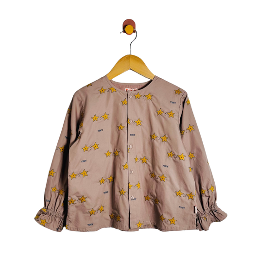 A children's long-sleeve button-front top in a muted taupe-brown color, decorated with an all-over print of yellow stars outlined in black. The top features elasticized cuffs and is shown hanging on a wooden hanger against a plain white background.