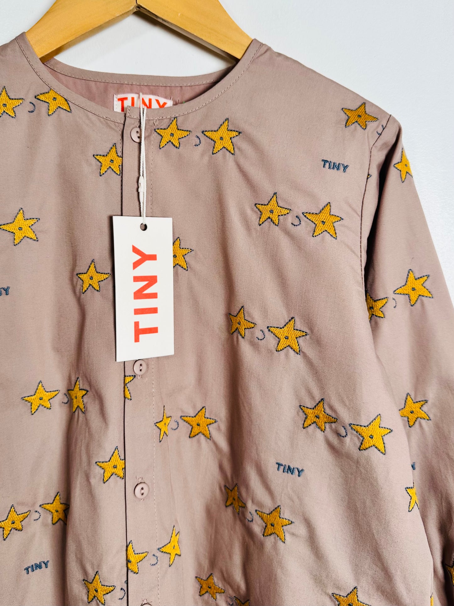 A close-up of a baby long-sleeve top in a dusty mauve color, featuring an all-over print of yellow stars with blue outlines. The top is displayed on a wooden hanger with a white 'TINY' brand tag hanging from the collar.