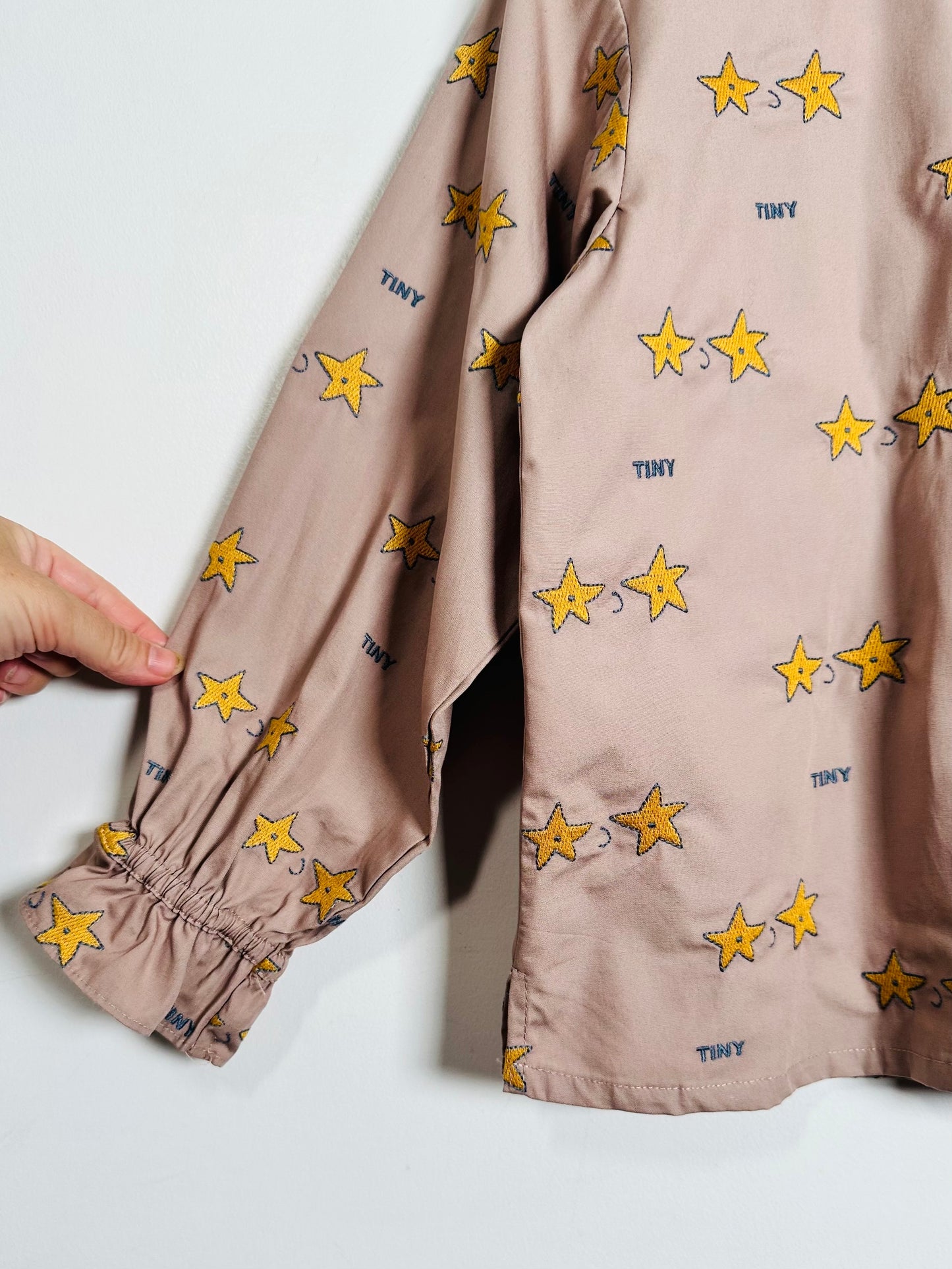 A close-up of a dusty pink long-sleeve shirt with an all-over print of yellow stars and small blue 'TINY' text, featuring a gathered elastic cuff on the sleeve. A hand is holding the cuff to demonstrate its stretch.