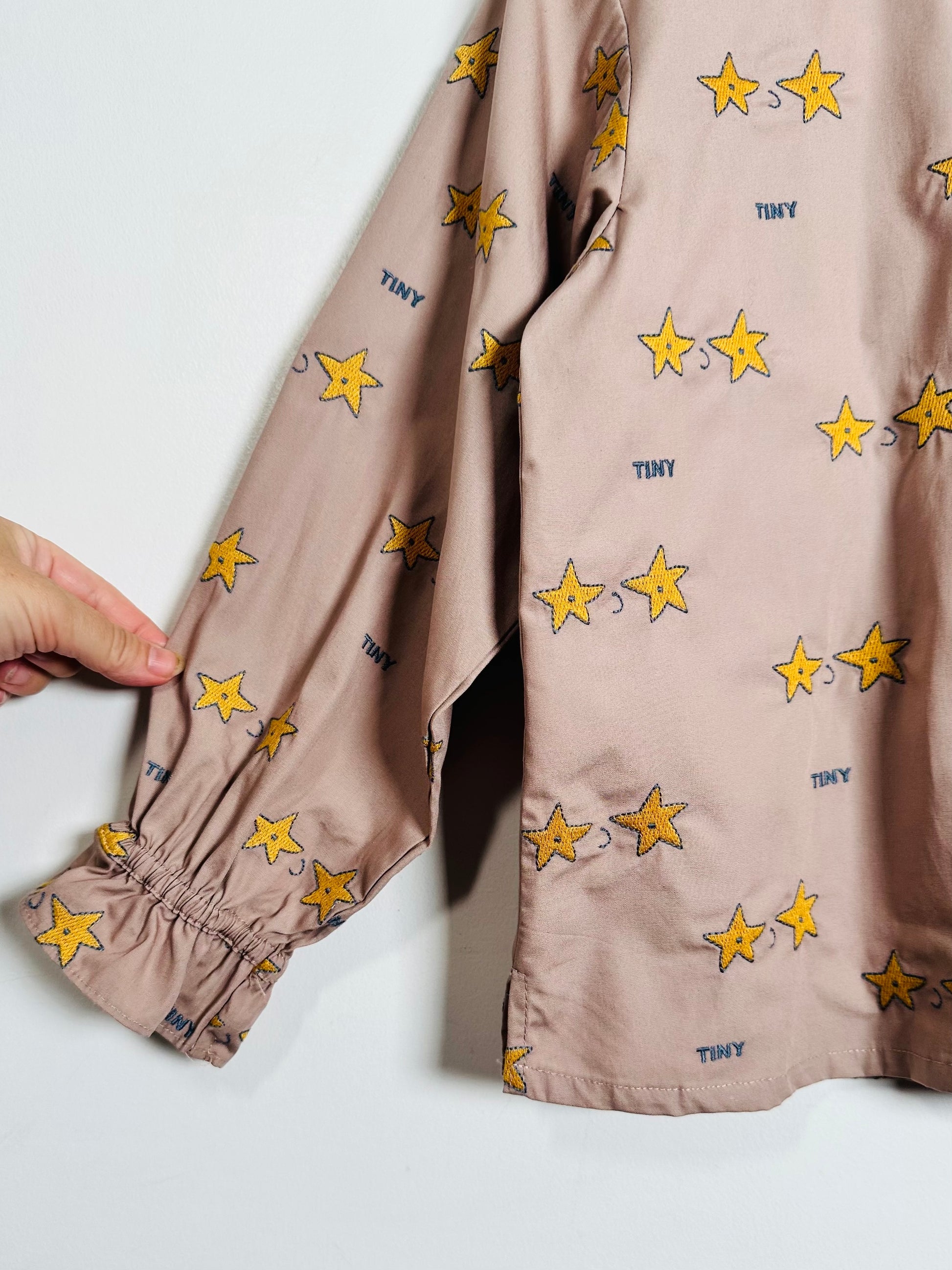 A close-up of a dusty pink long-sleeve shirt with an all-over print of yellow stars and small blue 'TINY' text, featuring a gathered elastic cuff on the sleeve. A hand is holding the cuff to demonstrate its stretch.