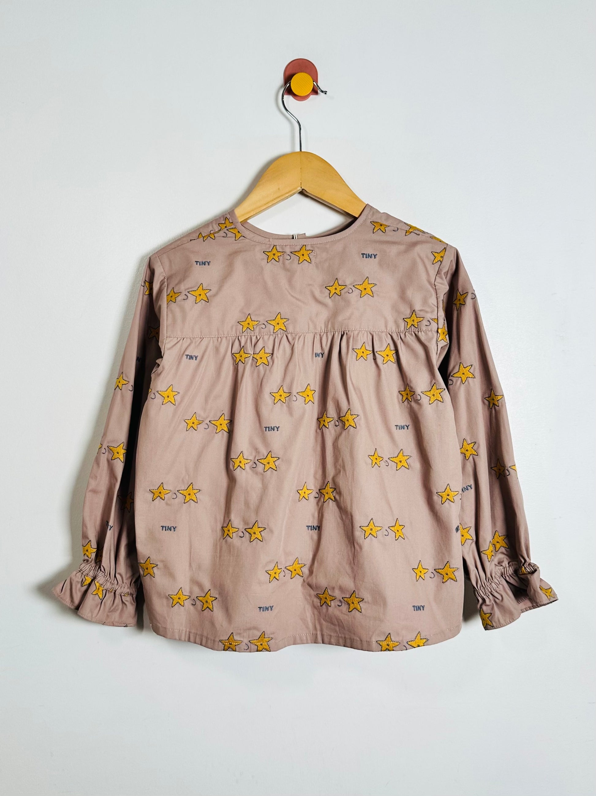 A long-sleeve babydoll top in dusty pink with an all-over yellow star print, hanging on a wooden hanger against a plain white wall.