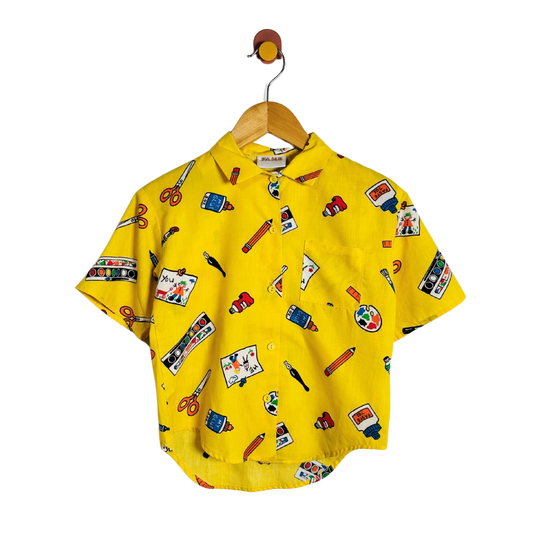 A yellow short-sleeve button-up shirt with an all-over colorful craft-themed print, hanging on a wooden hanger with a brown hook against a plain white background.
