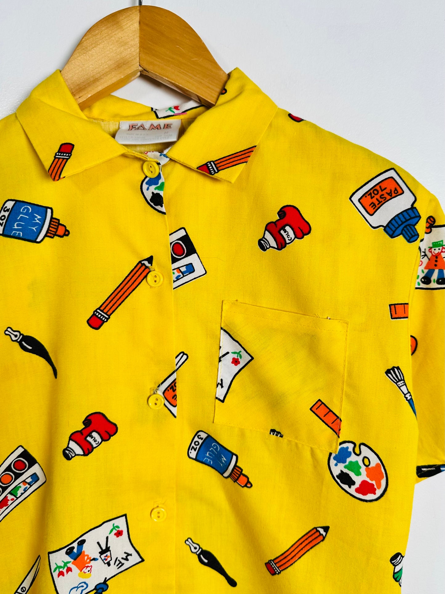 A bright yellow short-sleeve button-up shirt with an all-over colorful art-themed print, hanging on a wooden hanger against a plain light background.
