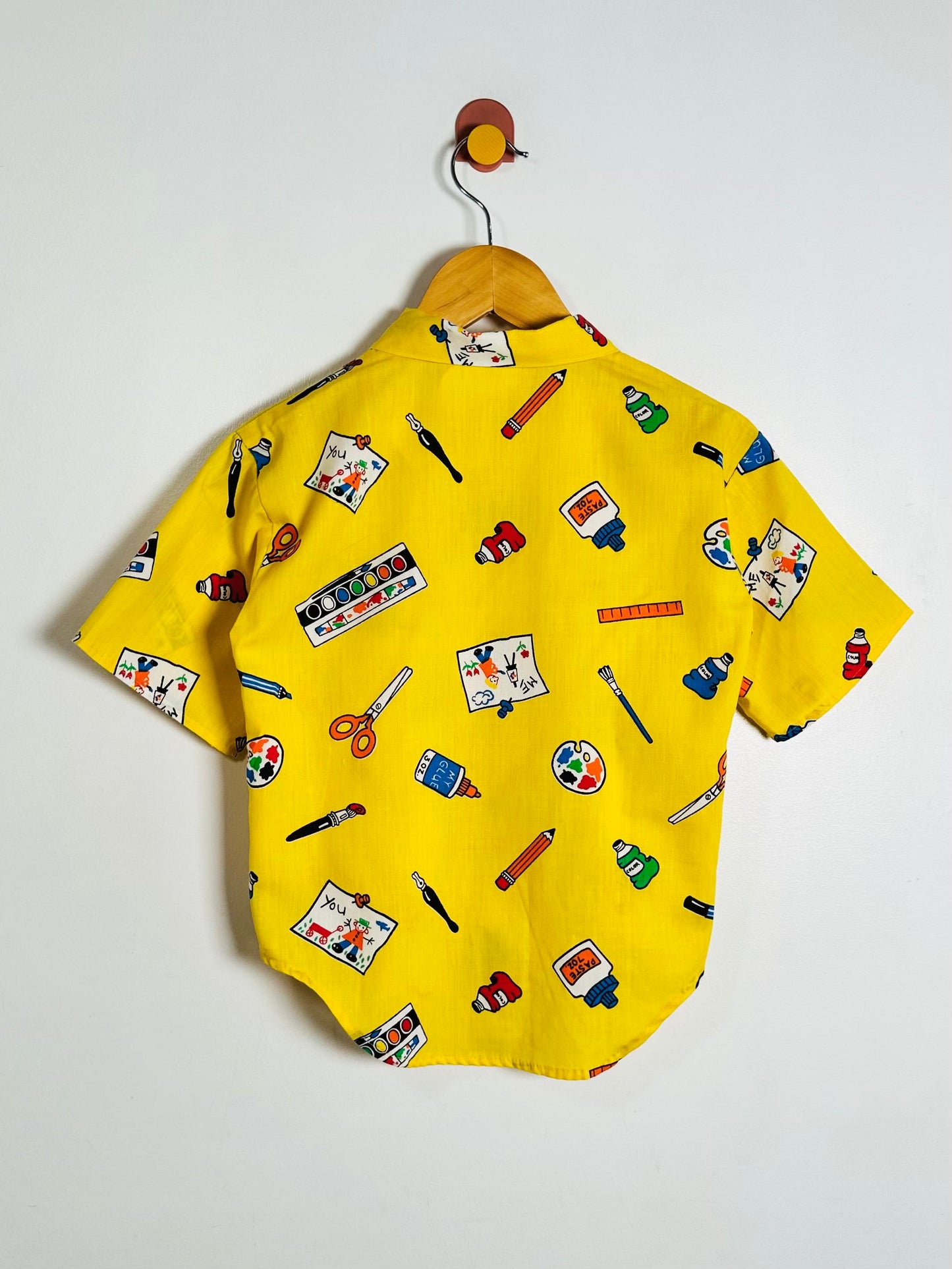 A yellow short-sleeve button-up shirt with an all-over colorful art-themed print, hanging on a wooden hanger against a plain white wall.
