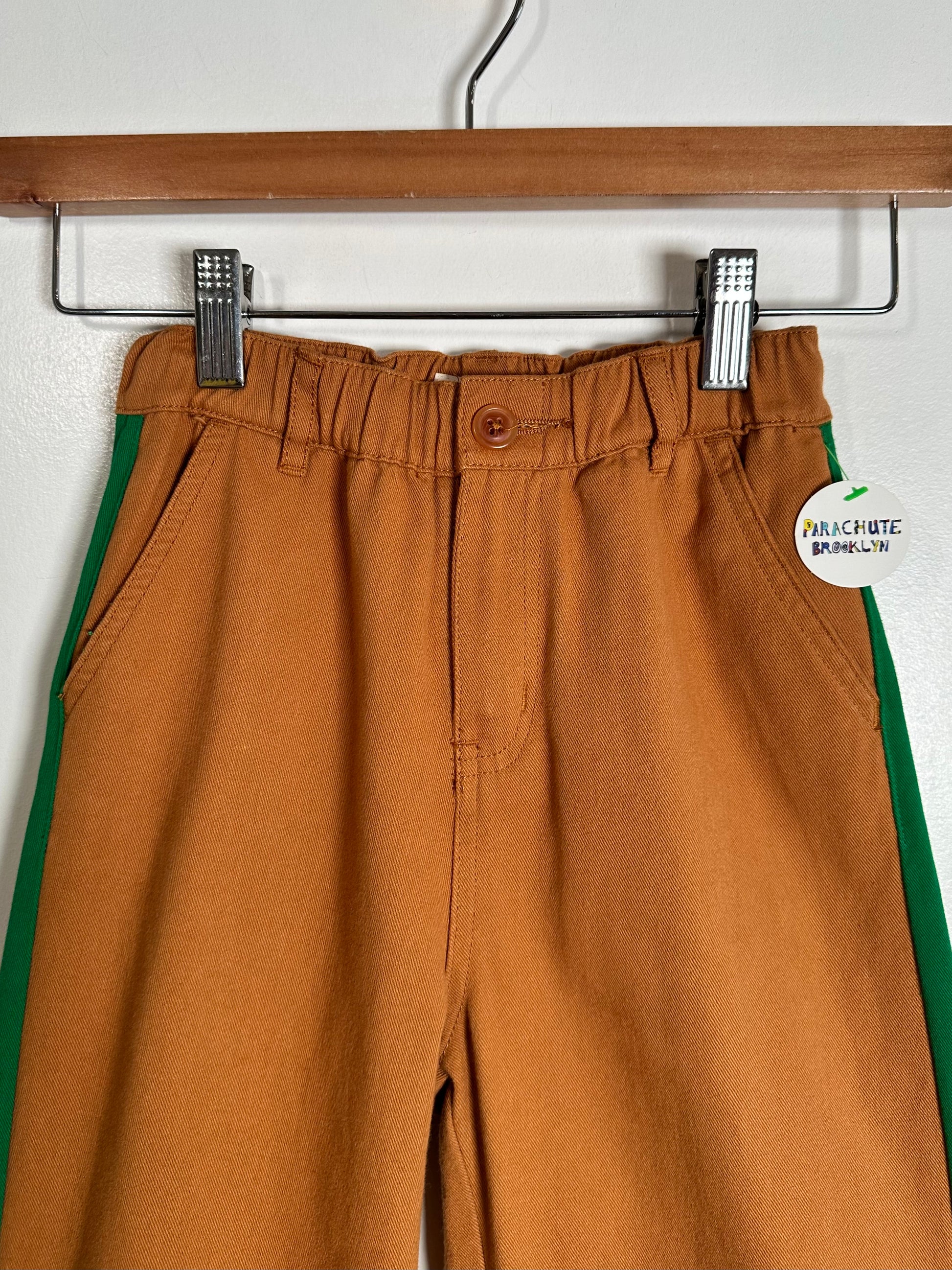 A pair of wide-leg trousers in a warm camel brown color, hanging on a wooden hanger. The pants feature a green stripe running down the side.