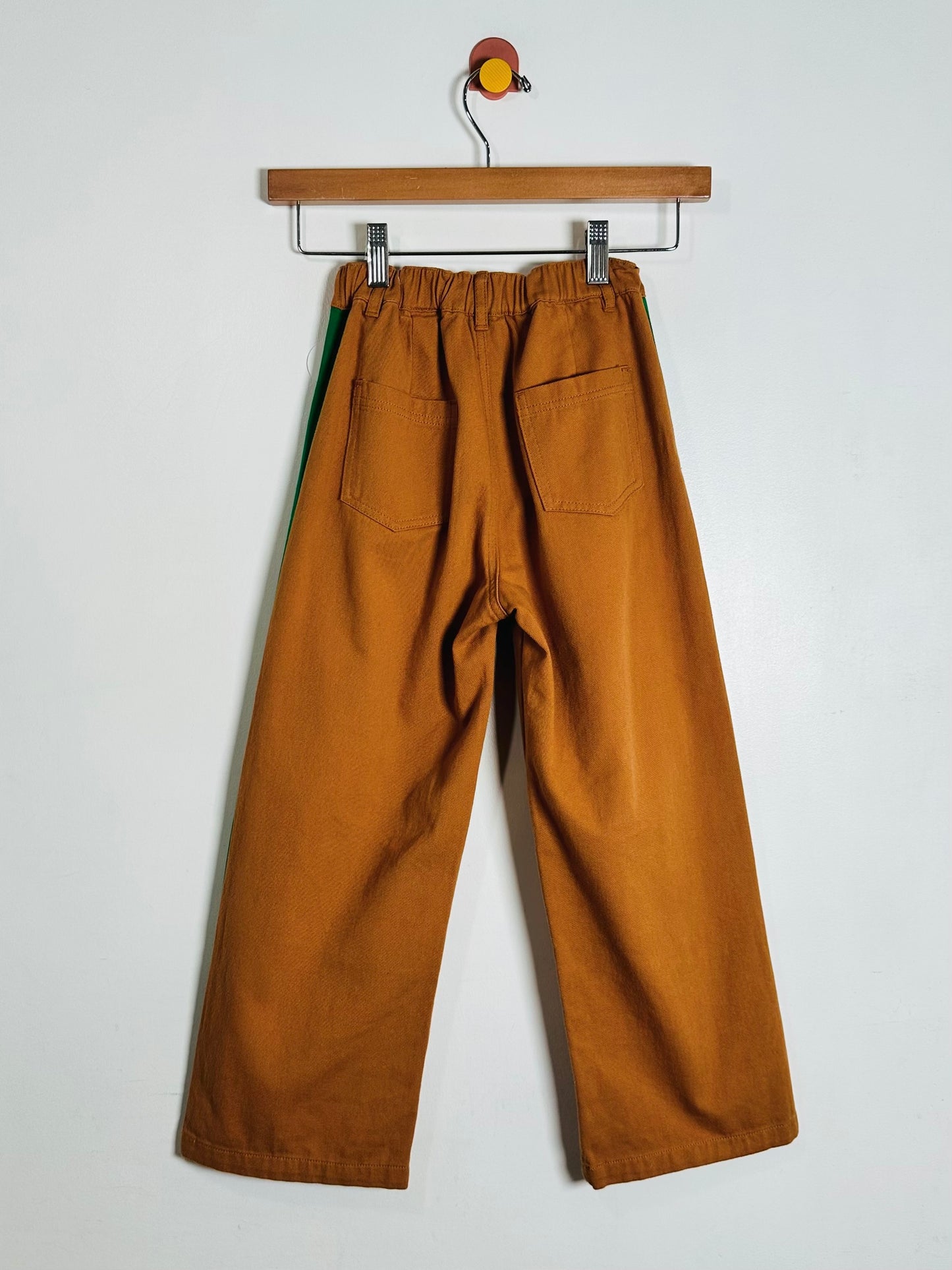 A pair of wide-leg brown pants with a green stripe detail on the side, hanging from a wooden hanger against a plain white wall.