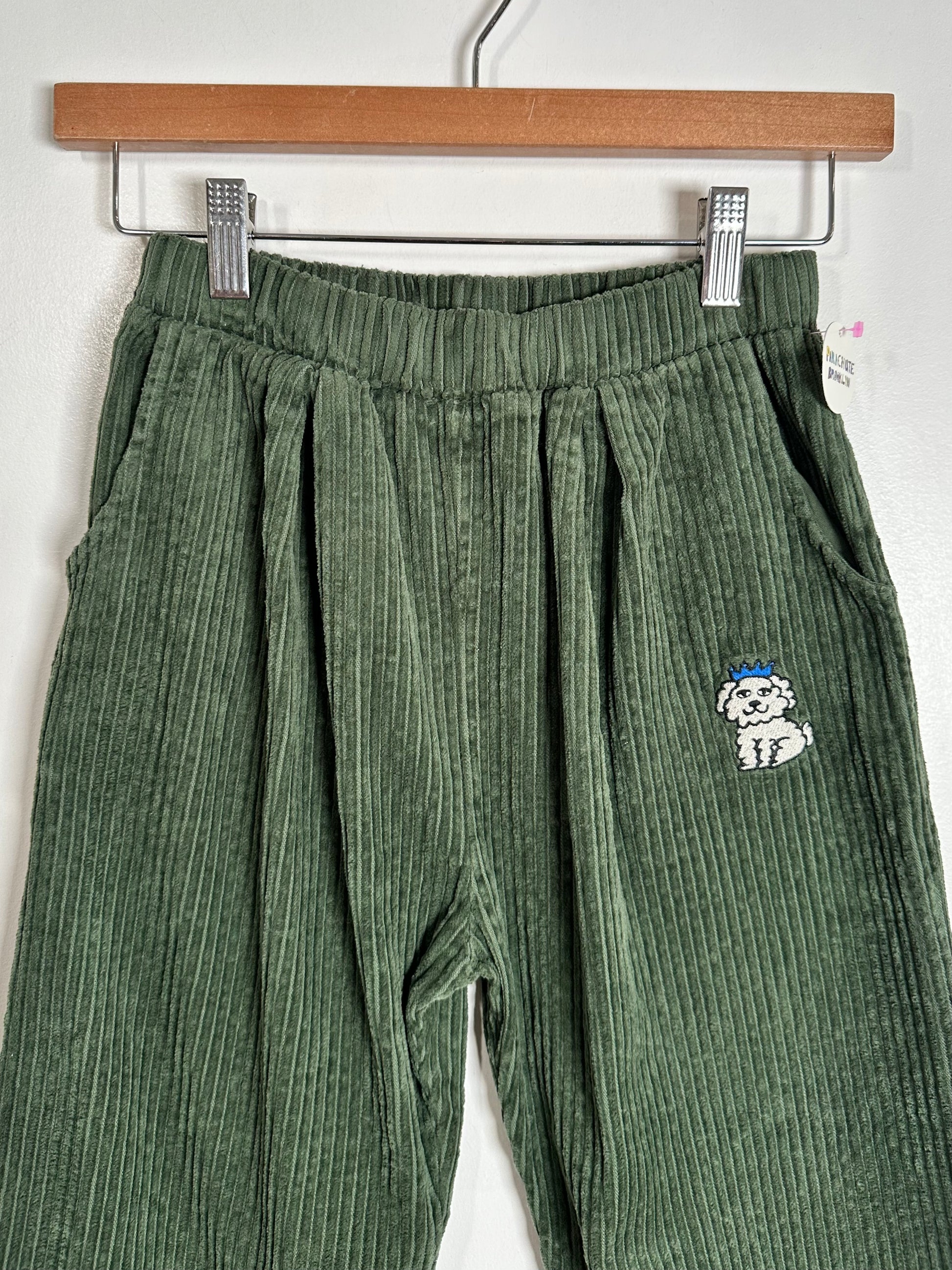 Green corduroy pants with an elastic waistband and a small embroidered dog patch on the left leg, hanging on a wooden hanger against a white wall.