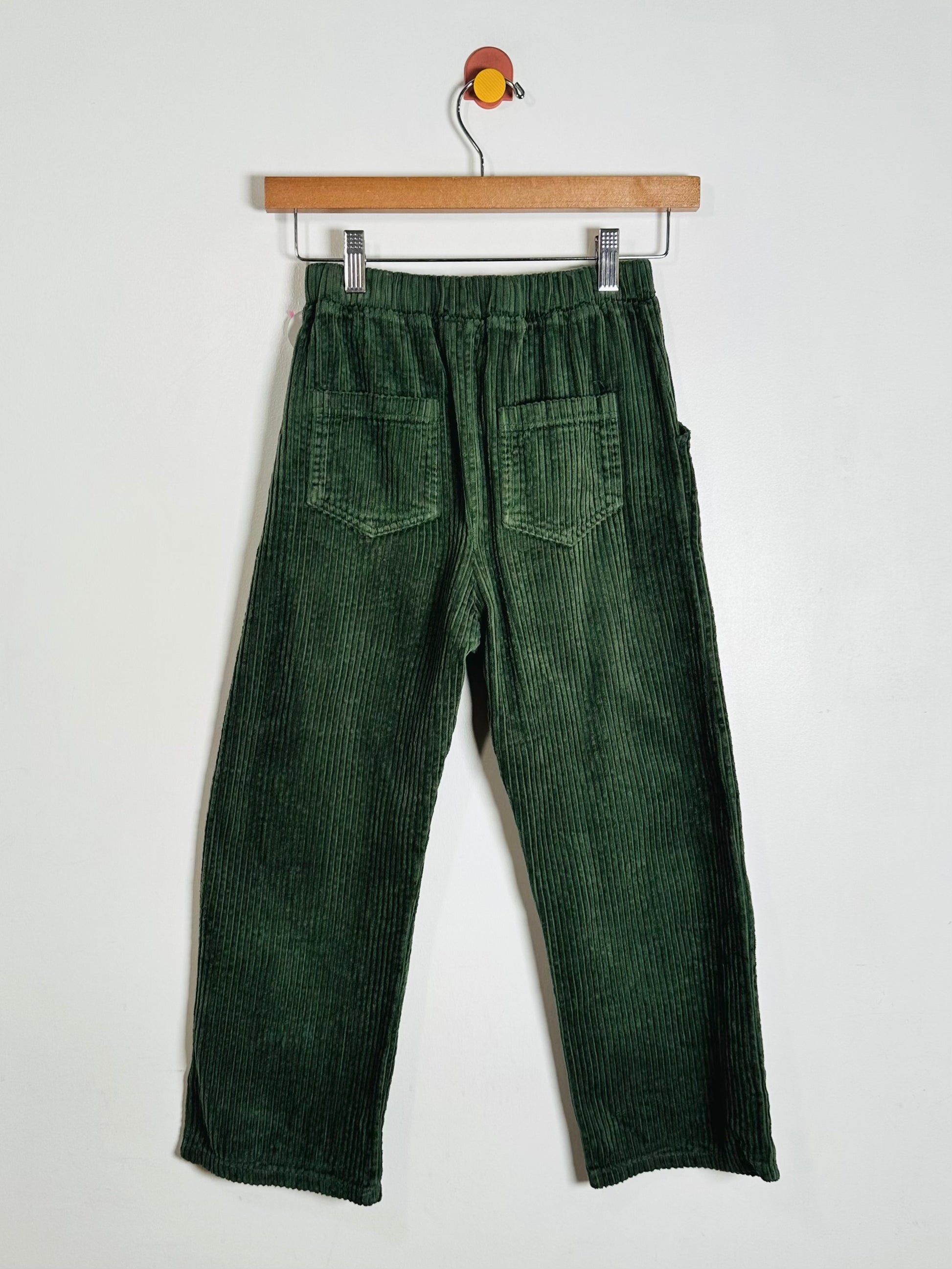 A pair of dark green corduroy pants hanging from a wooden hanger against a plain white wall.