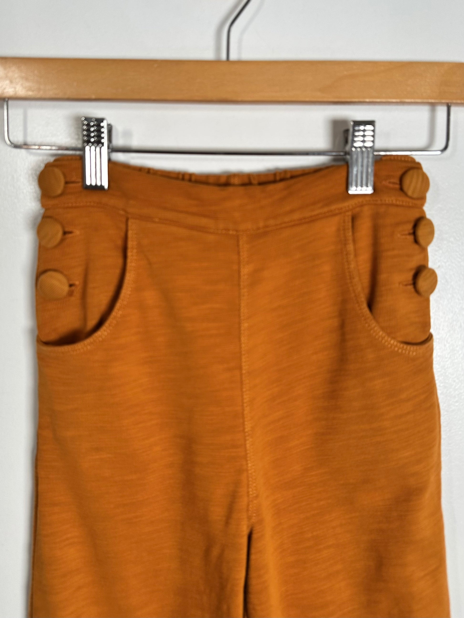A pair of mustard orange cropped pants with decorative button details on the waistband and side pockets, hanging from a wooden hanger with metal clips against a plain white wall.
