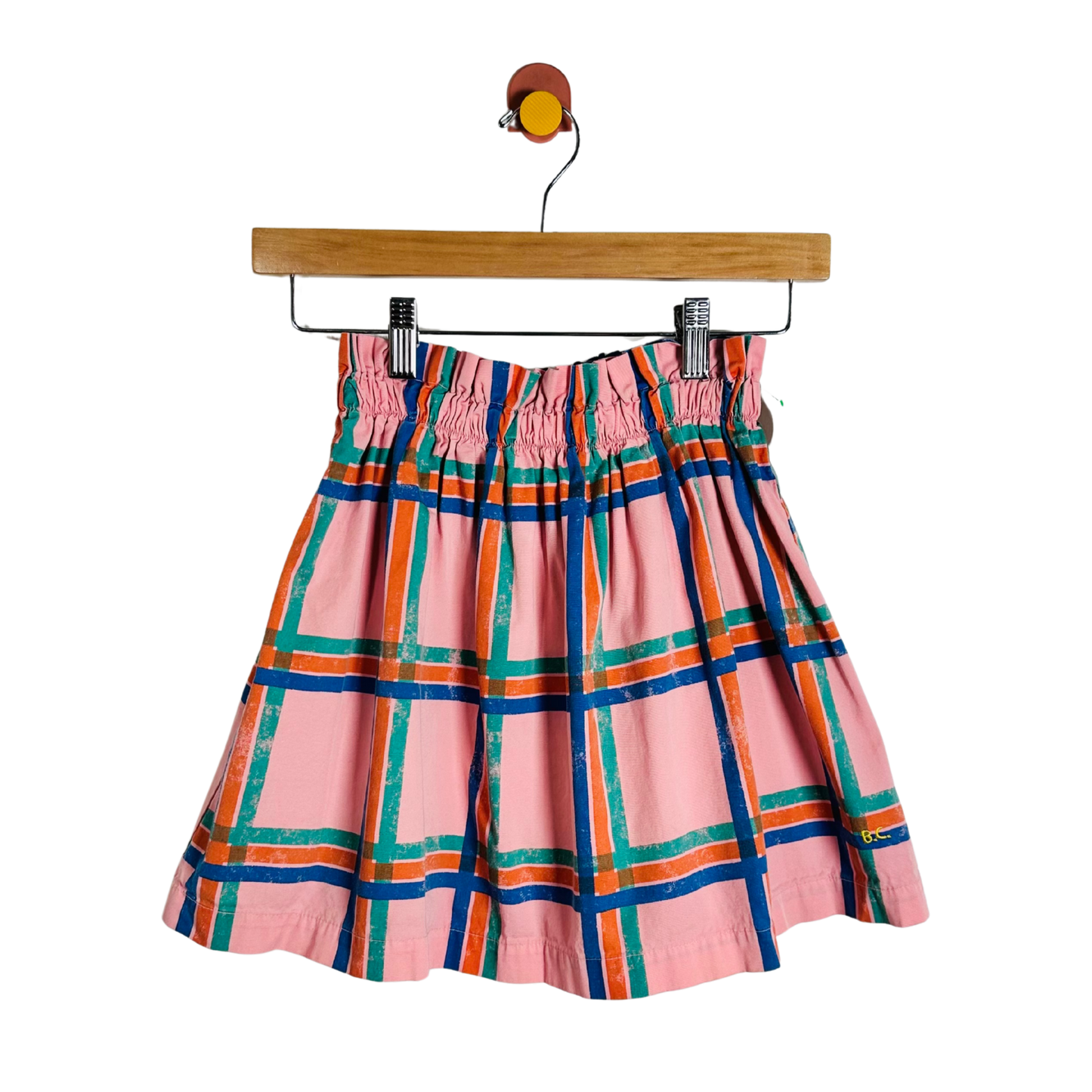 A pink pleated skirt with a colorful plaid pattern in blue, orange, and green, hanging from a wooden hanger against a white background.