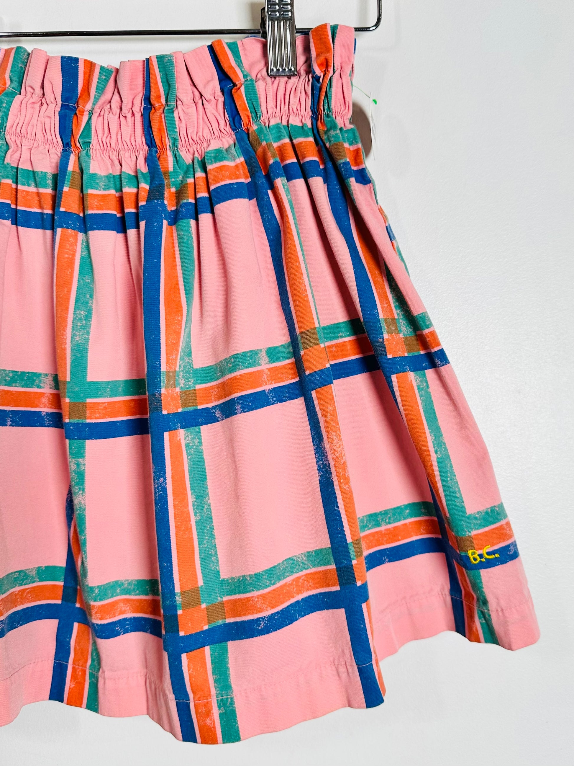 A pink pleated skirt with a colorful plaid pattern in blue, orange, and green, hanging on a black hanger against a plain white wall.