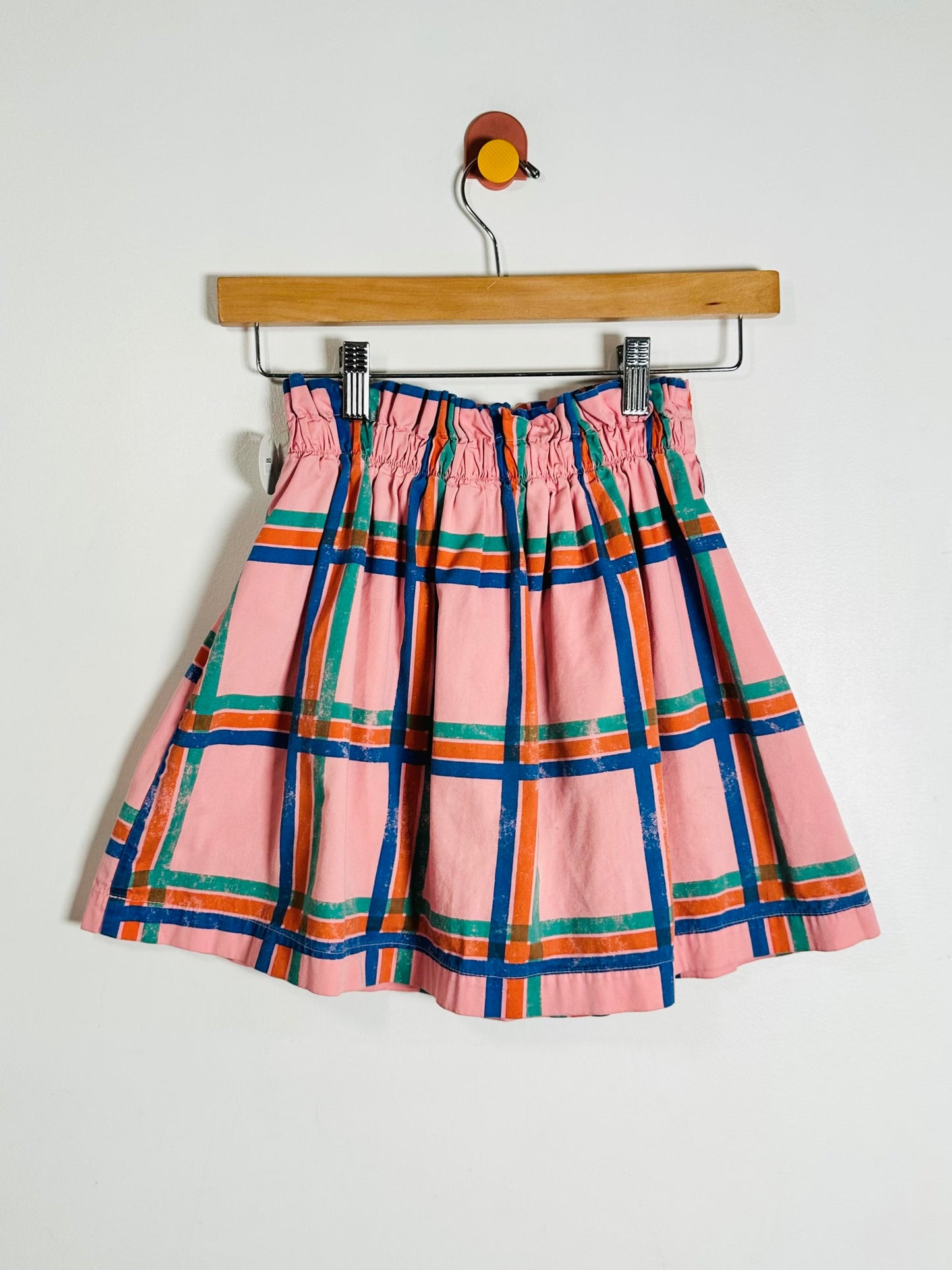 A pink plaid skirt with blue, green, and orange stripes, hanging from a wooden hanger against a plain white wall.