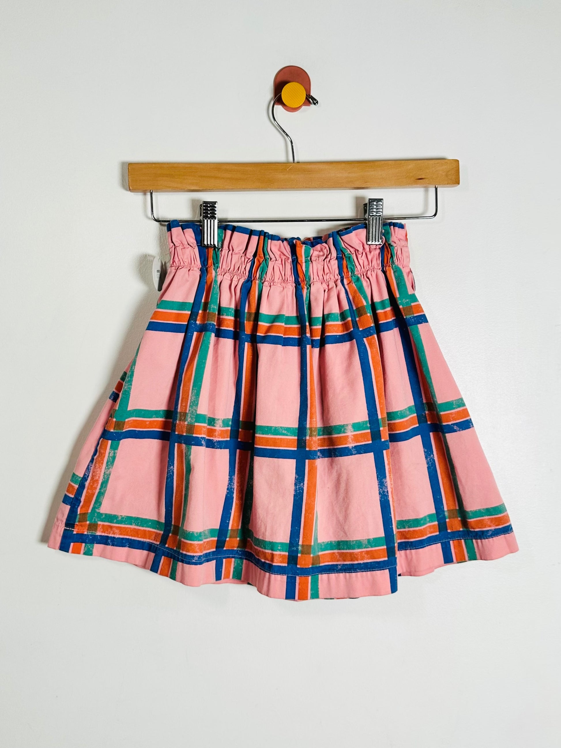 A pink plaid skirt with blue, green, and orange stripes, hanging from a wooden hanger against a plain white wall.