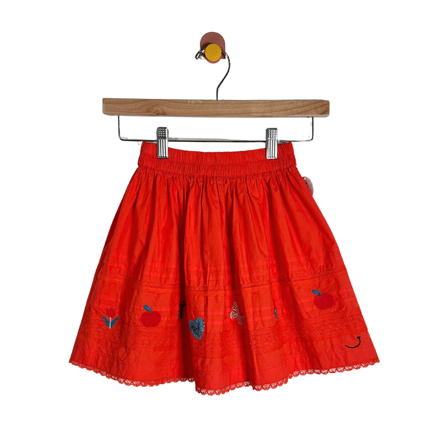 A bright red cotton skirt with embroidered apple motifs and a scalloped lace hem, hanging from a wooden hanger against a plain white background.