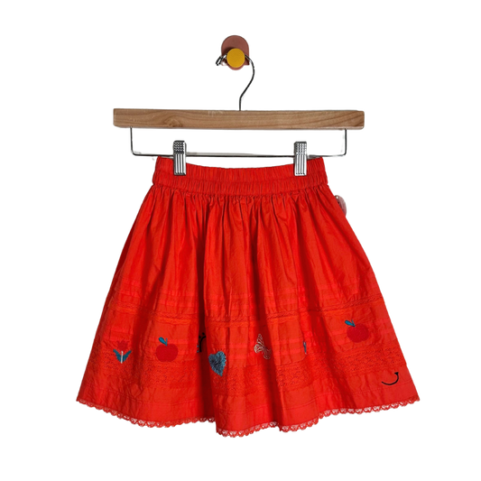 A bright red cotton skirt with embroidered apple motifs and a scalloped lace hem, hanging from a wooden hanger against a plain white background.