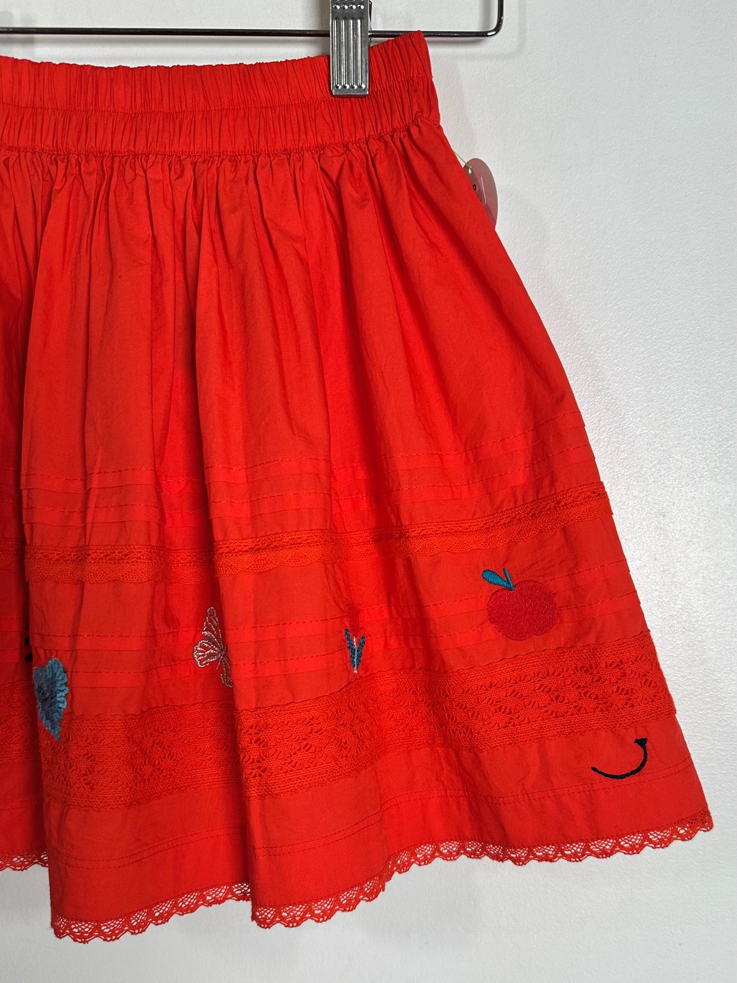 A bright red cotton skirt with embroidered details, including a red apple with a blue leaf, a butterfly, and a smiley face, hanging on a metal hanger against a white wall.