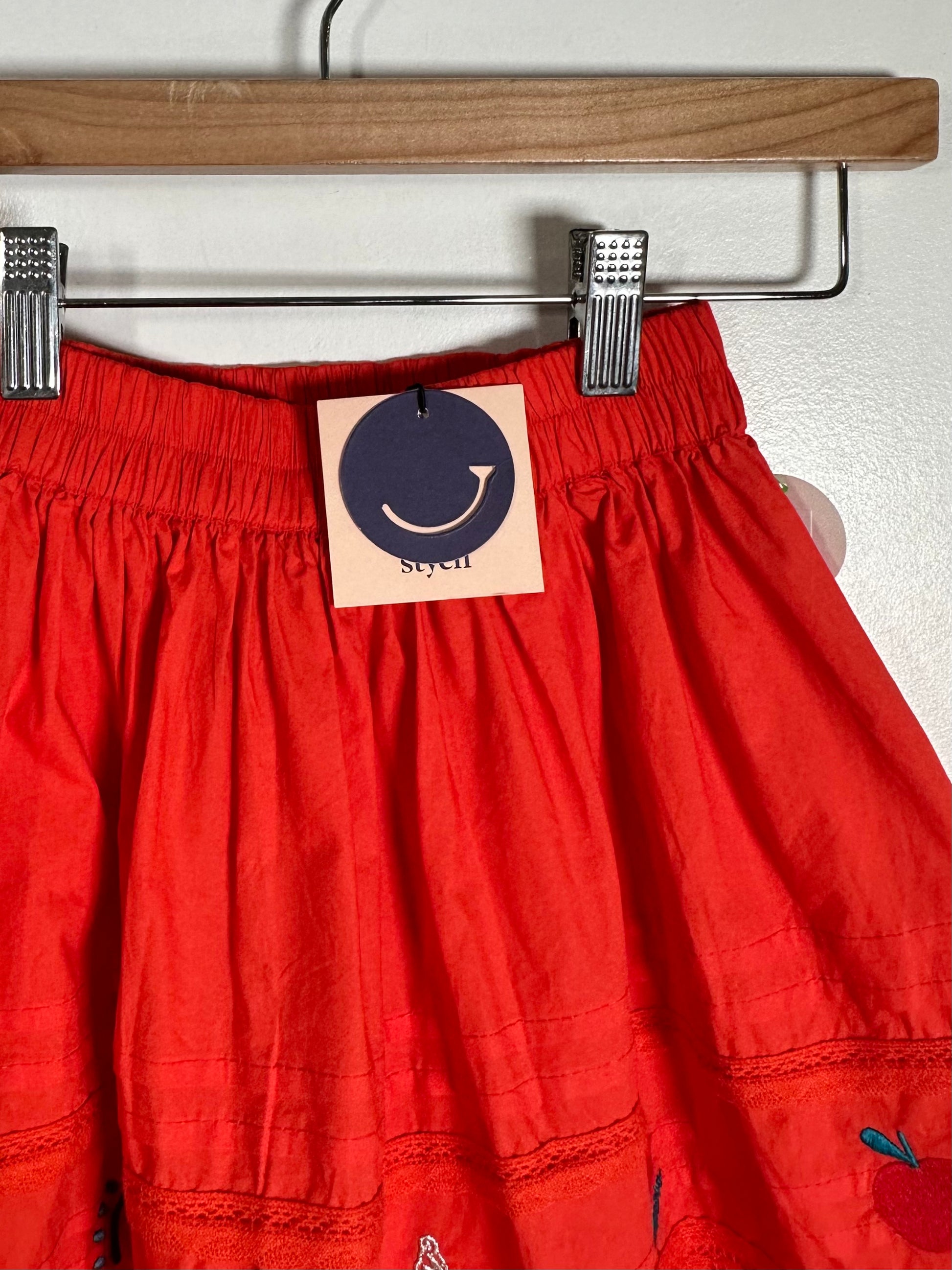 A close-up of a bright red skirt hanging from a wooden hanger with metal clips. A paper tag with a smiley face logo and the brand name 'syeh' is attached at the waistband.