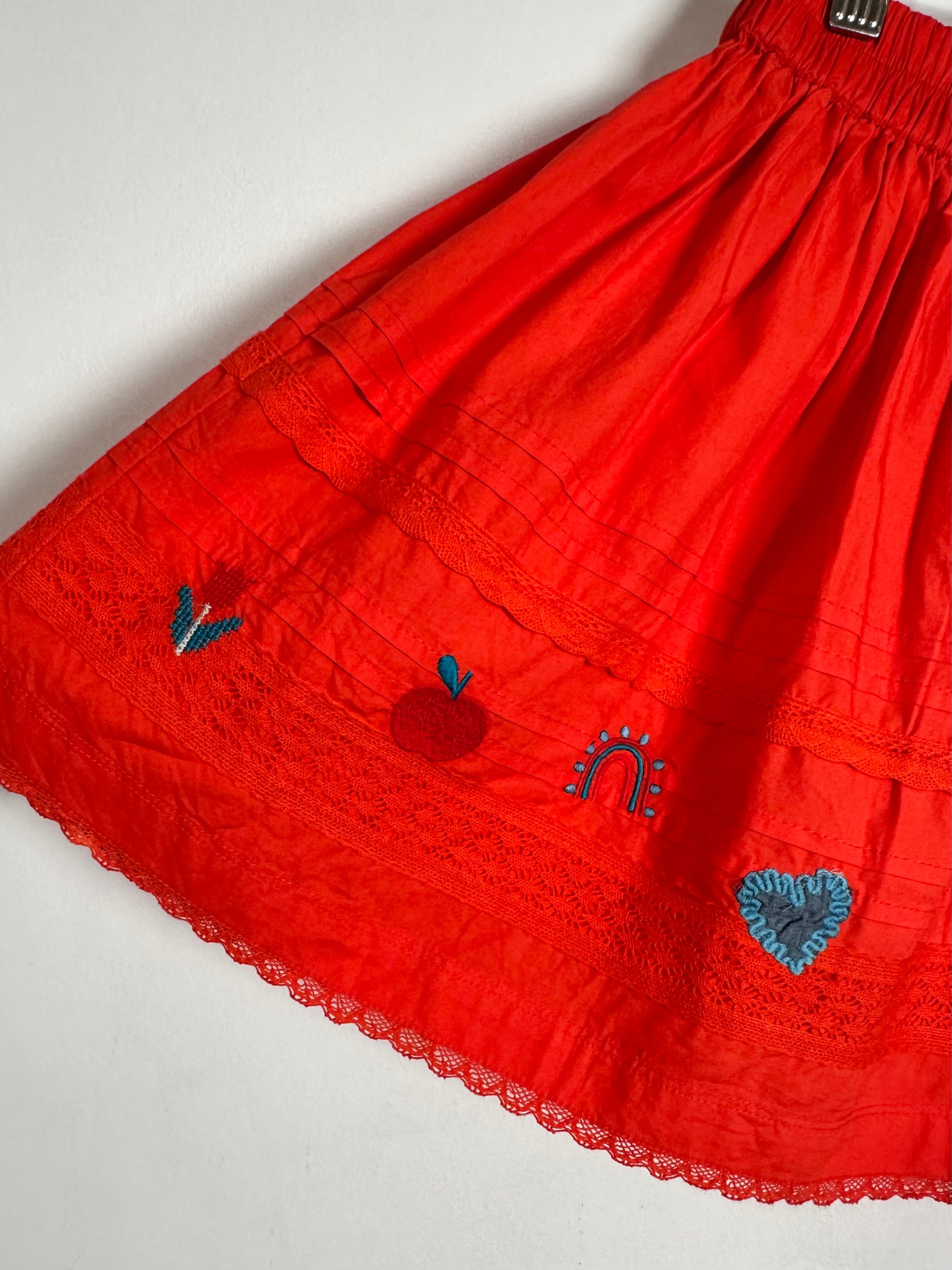 A bright red cotton skirt with embroidered motifs including a blue tulip, red apple, rainbow, and blue heart, finished with a lace hem.