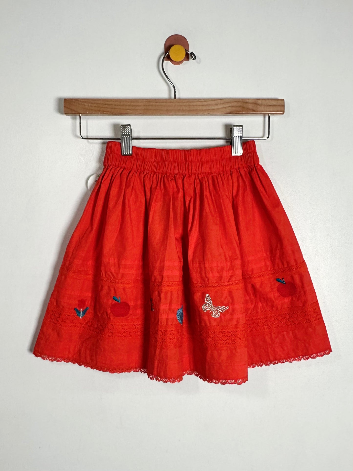 A bright red cotton skirt with embroidered details, hanging on a wooden hanger against a plain white wall.