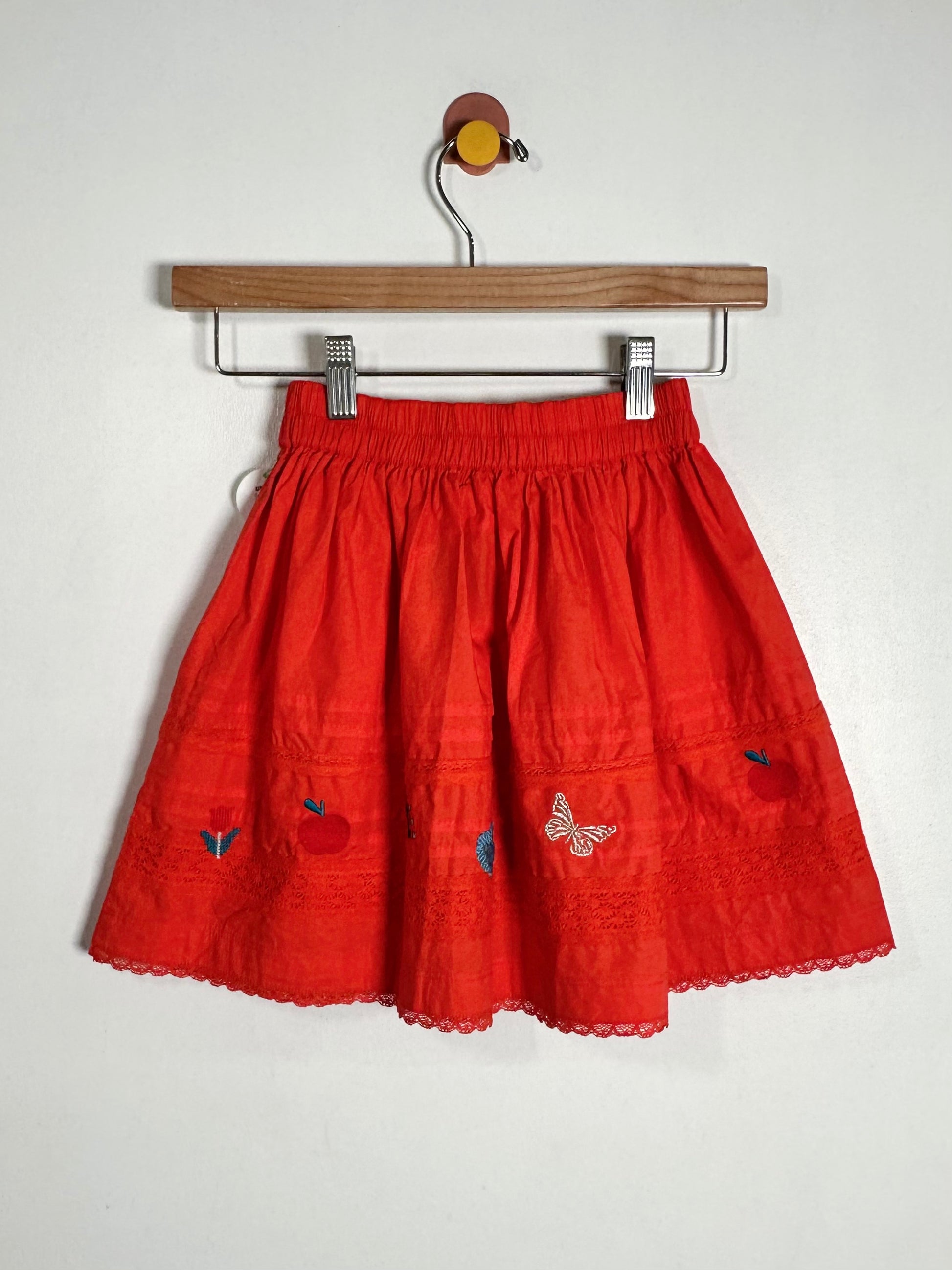 A bright red cotton skirt with embroidered details, hanging on a wooden hanger against a plain white wall.