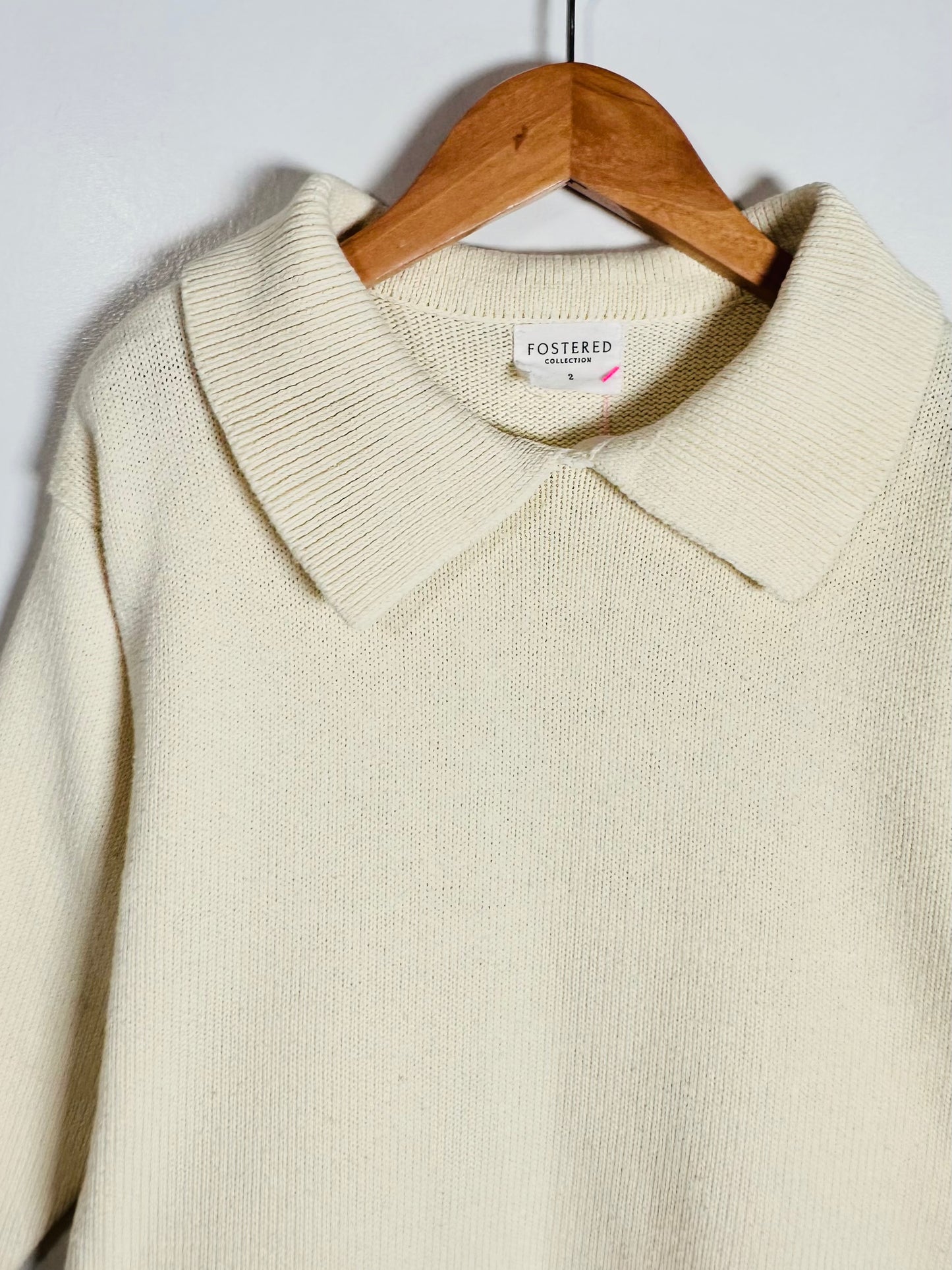 Cream-colored knit polo sweater hanging on a wooden hanger against a plain light gray wall. The brand label 'FOSTERED' is visible inside the collar.