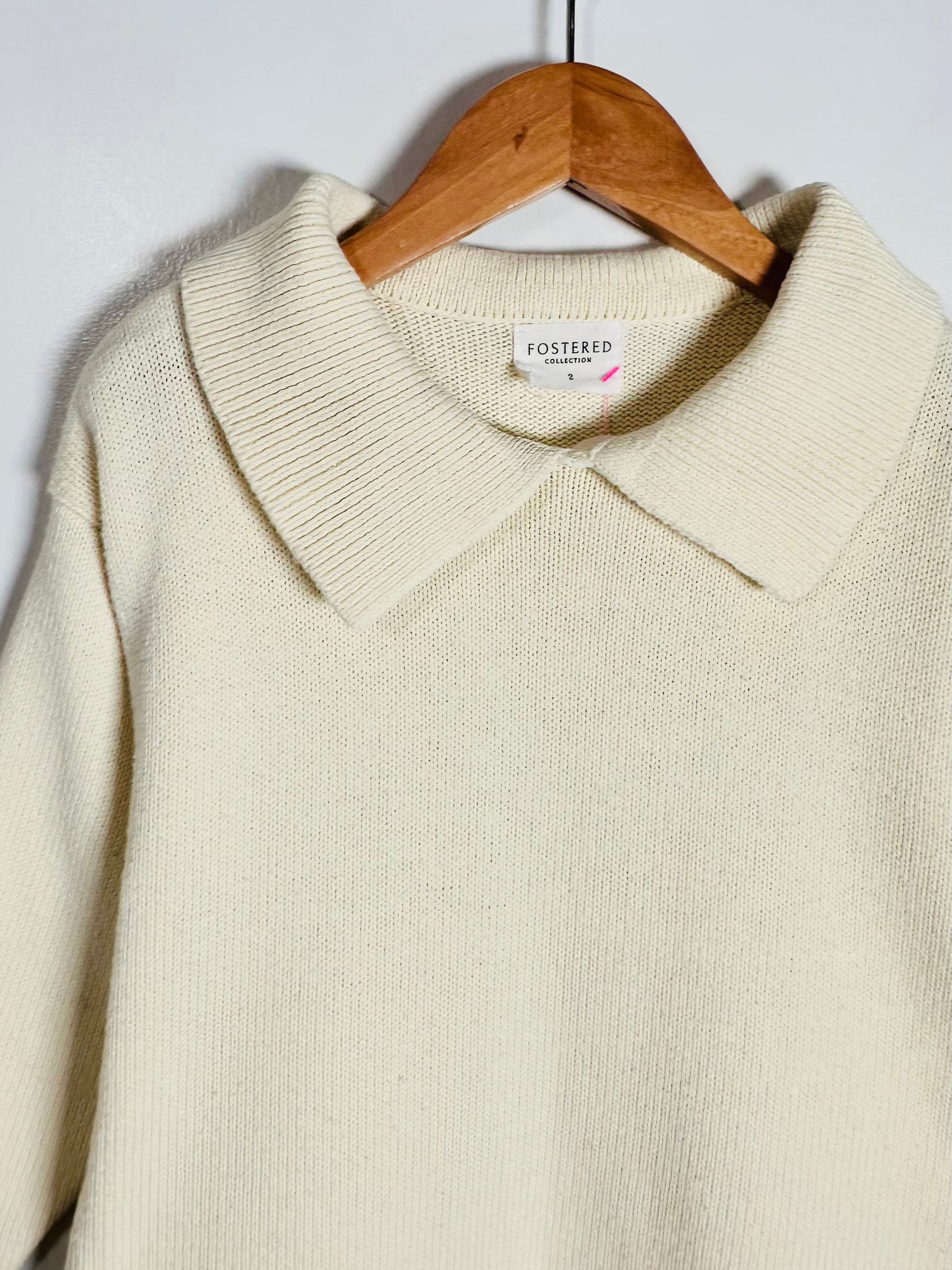 Cream-colored knit polo sweater hanging on a wooden hanger against a plain light gray wall. The brand label 'FOSTERED' is visible inside the collar.