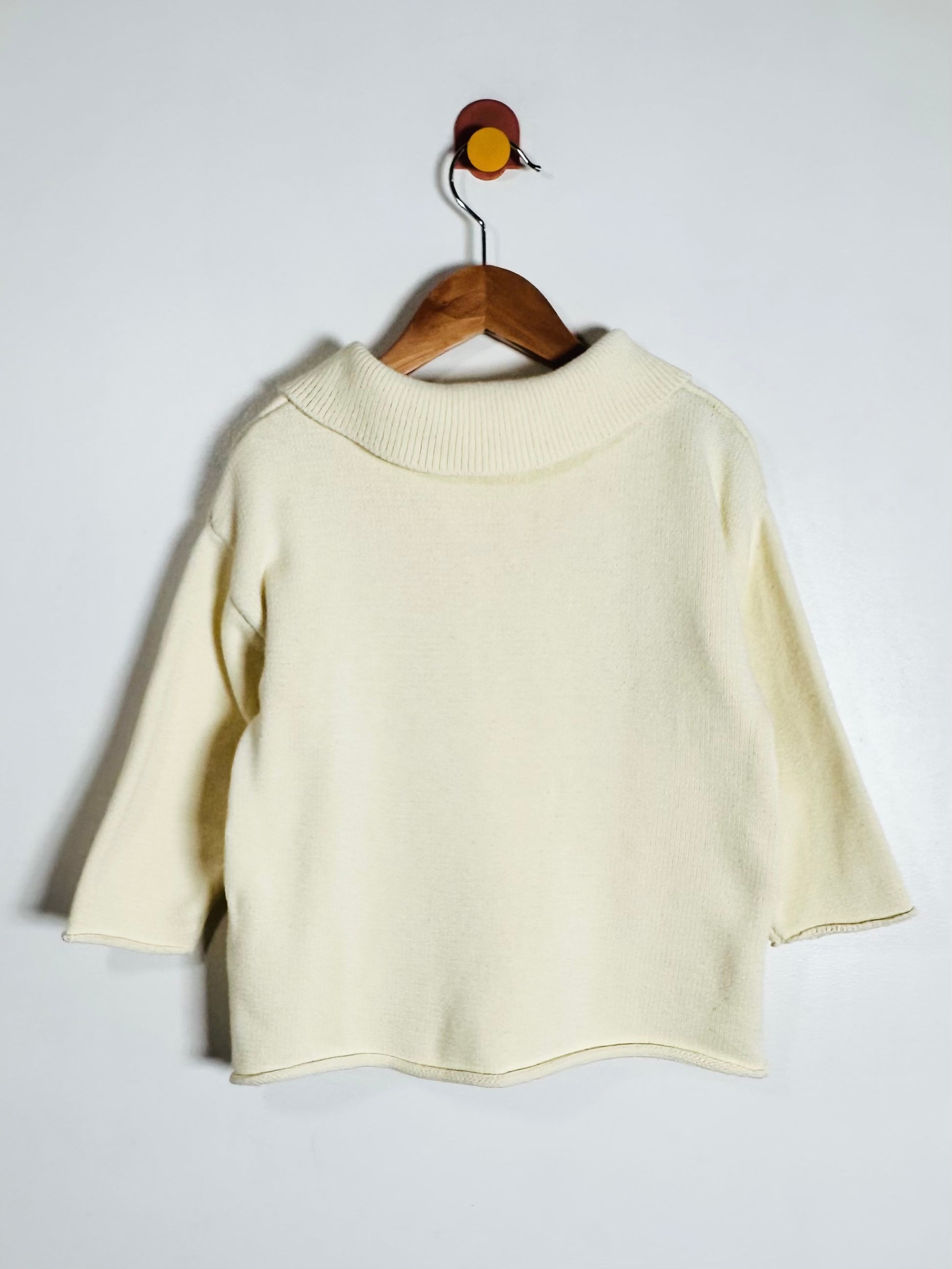 A cream-colored knit sweater with three-quarter sleeves and a ribbed mock neck, hanging on a wooden hanger against a plain light gray wall.