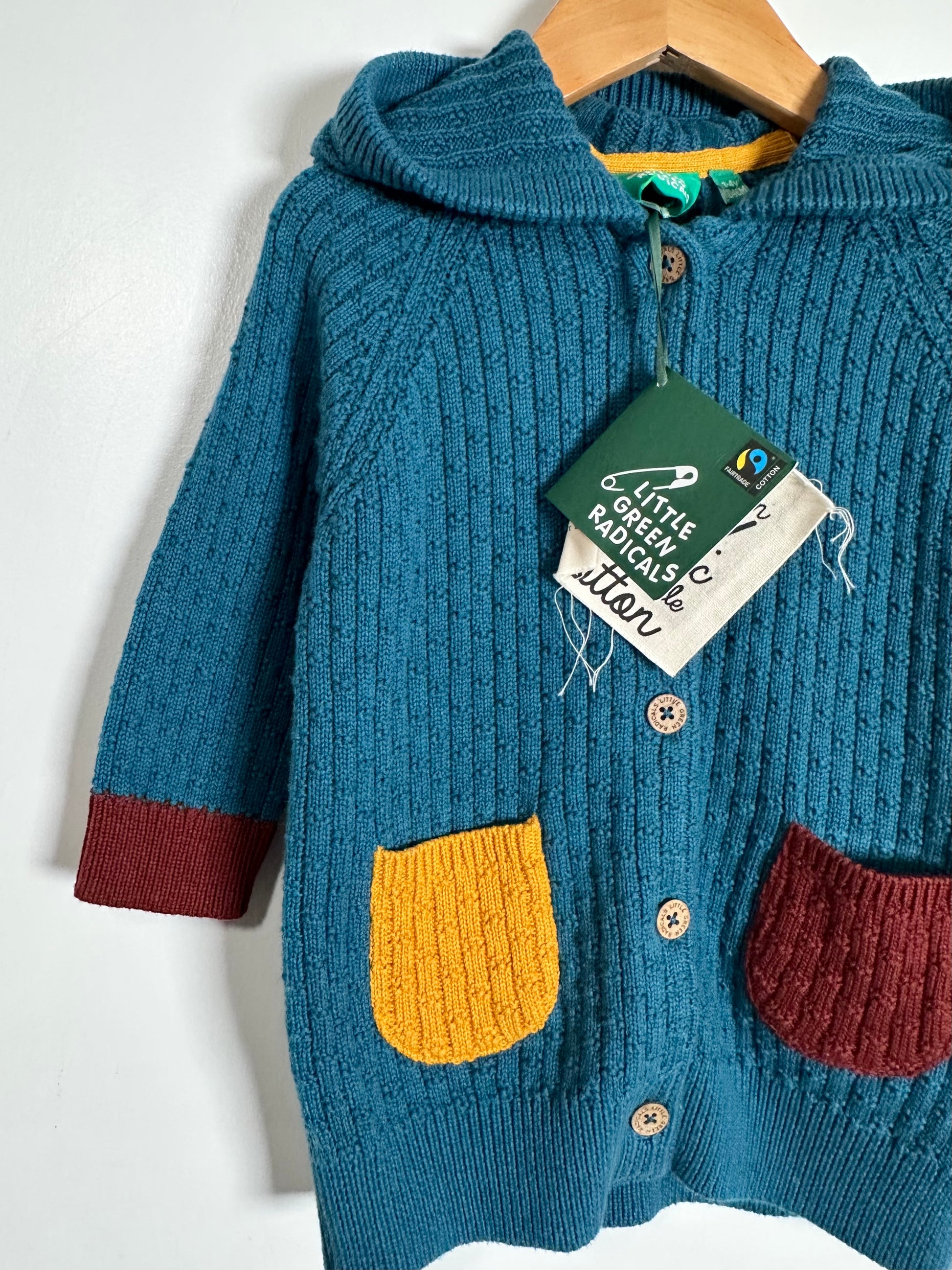 A teal blue ribbed knit hooded cardigan sweater with yellow and burgundy patch pockets, hanging on a wooden hanger with a 'Gentle Monster' brand tag attached.