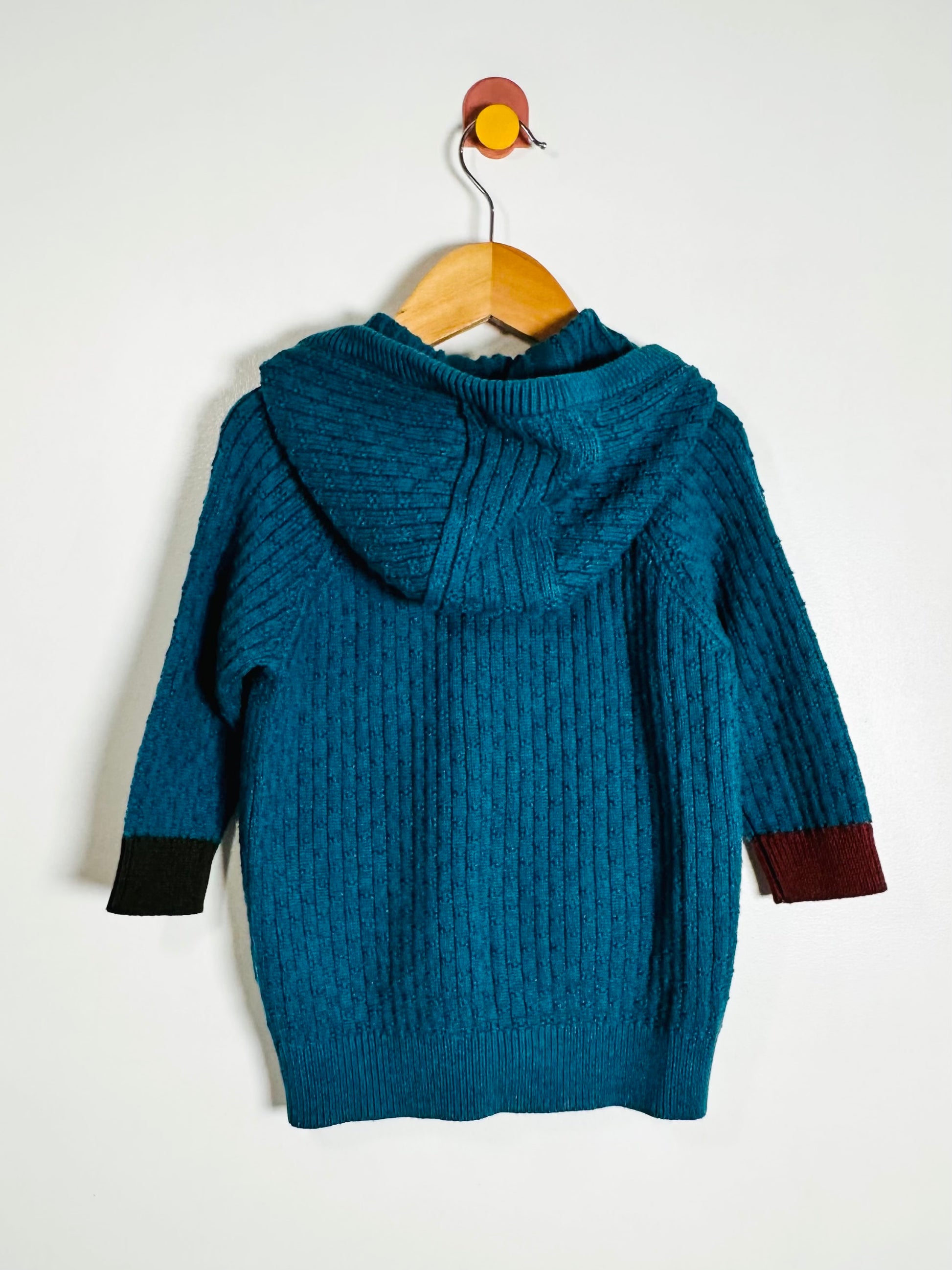 A teal blue cable-knit hooded sweater hanging on a wooden hanger against a plain white wall. The sweater features contrasting dark brown cuffs on one side and dark black cuffs on the other.