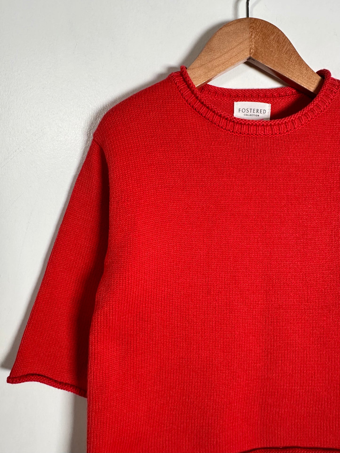 A bright red knit short-sleeve sweater hanging on a wooden hanger against a plain light gray wall. A small neck label reads FOSTERO.