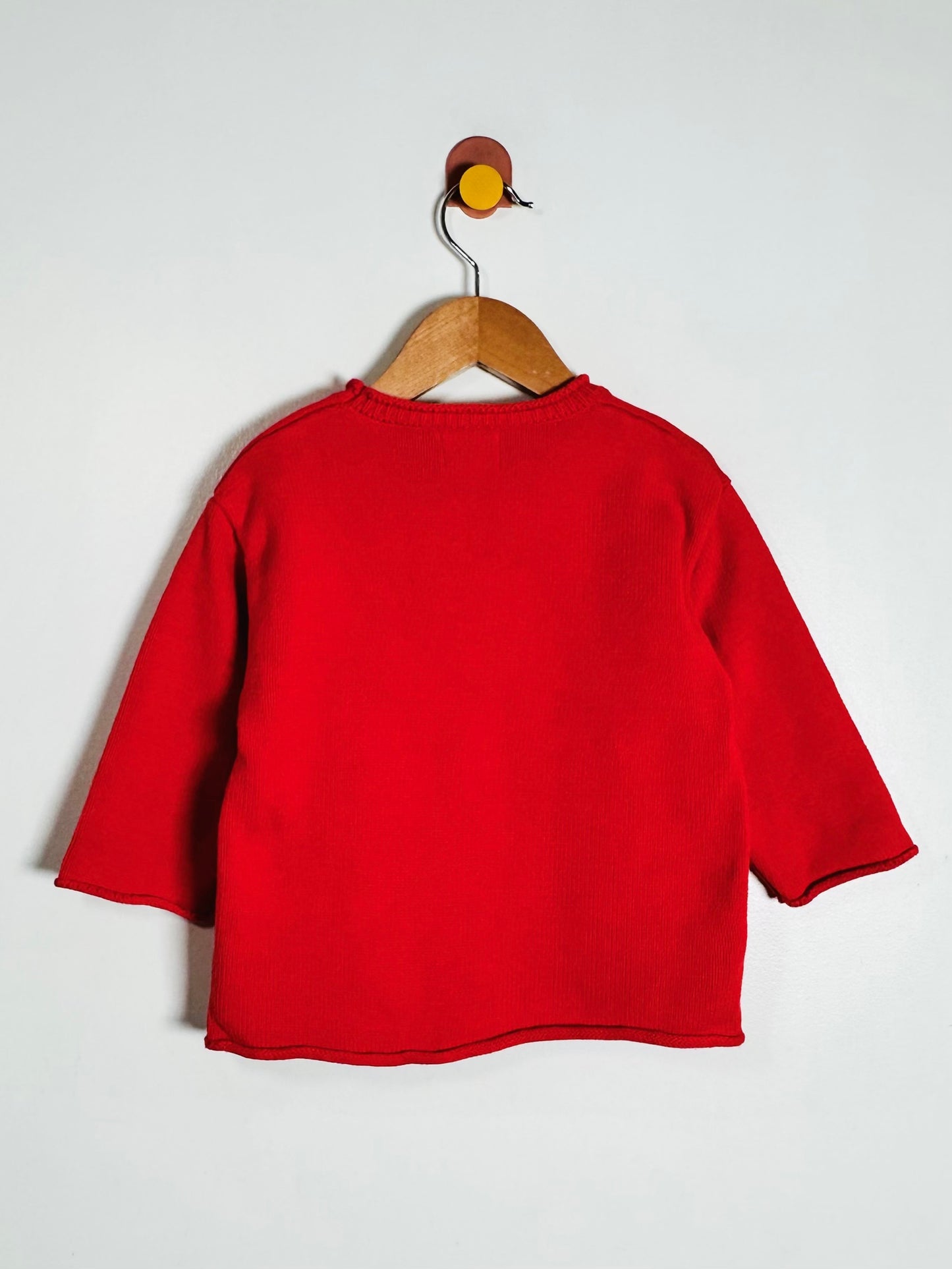 A bright red long-sleeve top hanging on a wooden hanger against a plain light gray wall.