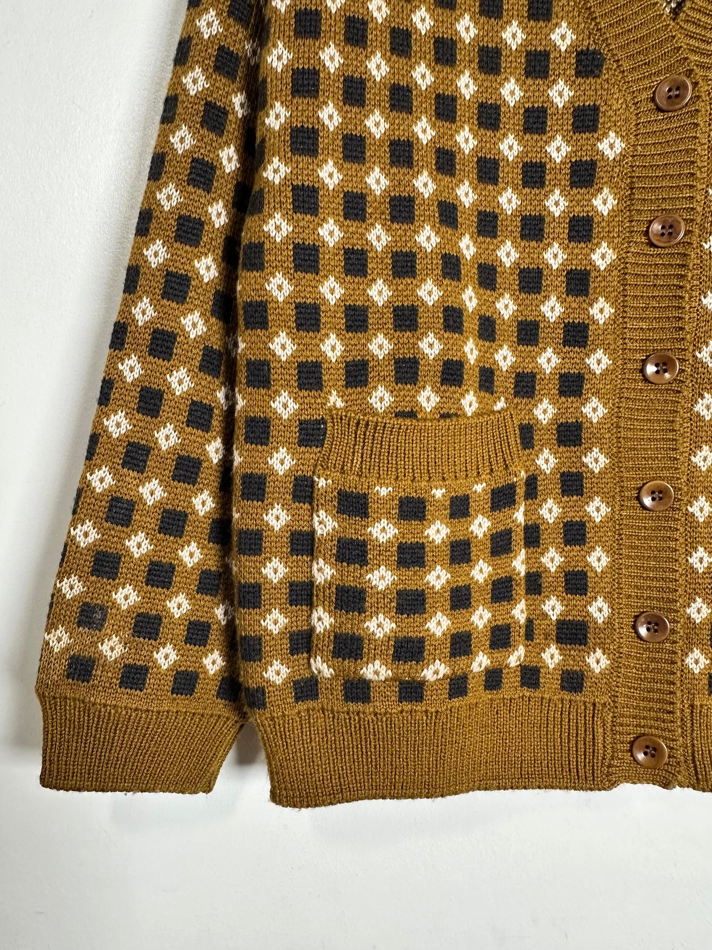 A close-up of a brown knit cardigan sweater featuring a black and cream geometric diamond pattern, a front pocket, and brown wooden buttons.
