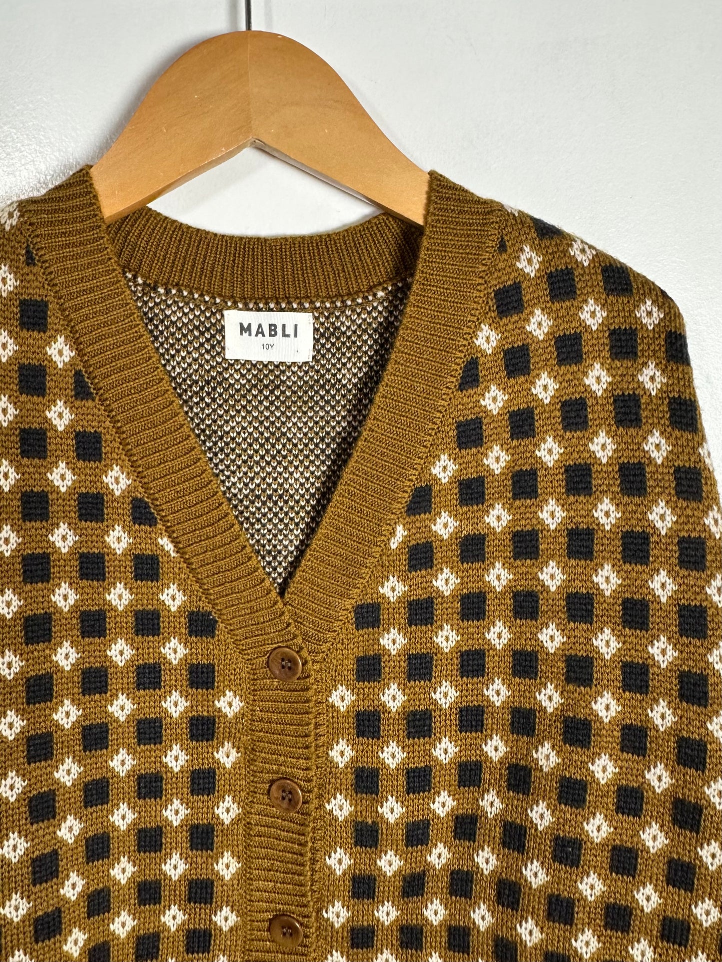A close-up of a MABLI knit cardigan sweater hanging on a wooden hanger against a light gray wall. The cardigan features a mustard brown base with a black and white geometric diamond pattern.