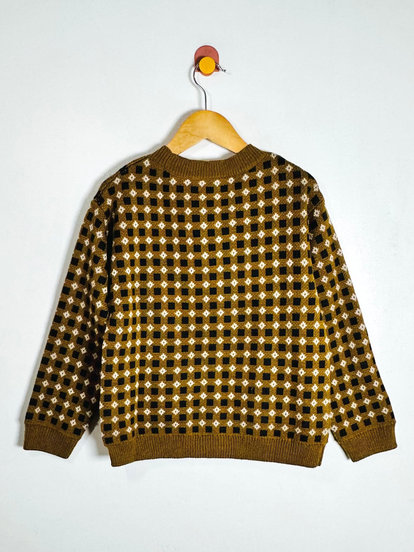 A long-sleeve knit sweater with a brown, black, and white geometric pattern, hanging on a wooden hanger against a plain white wall.