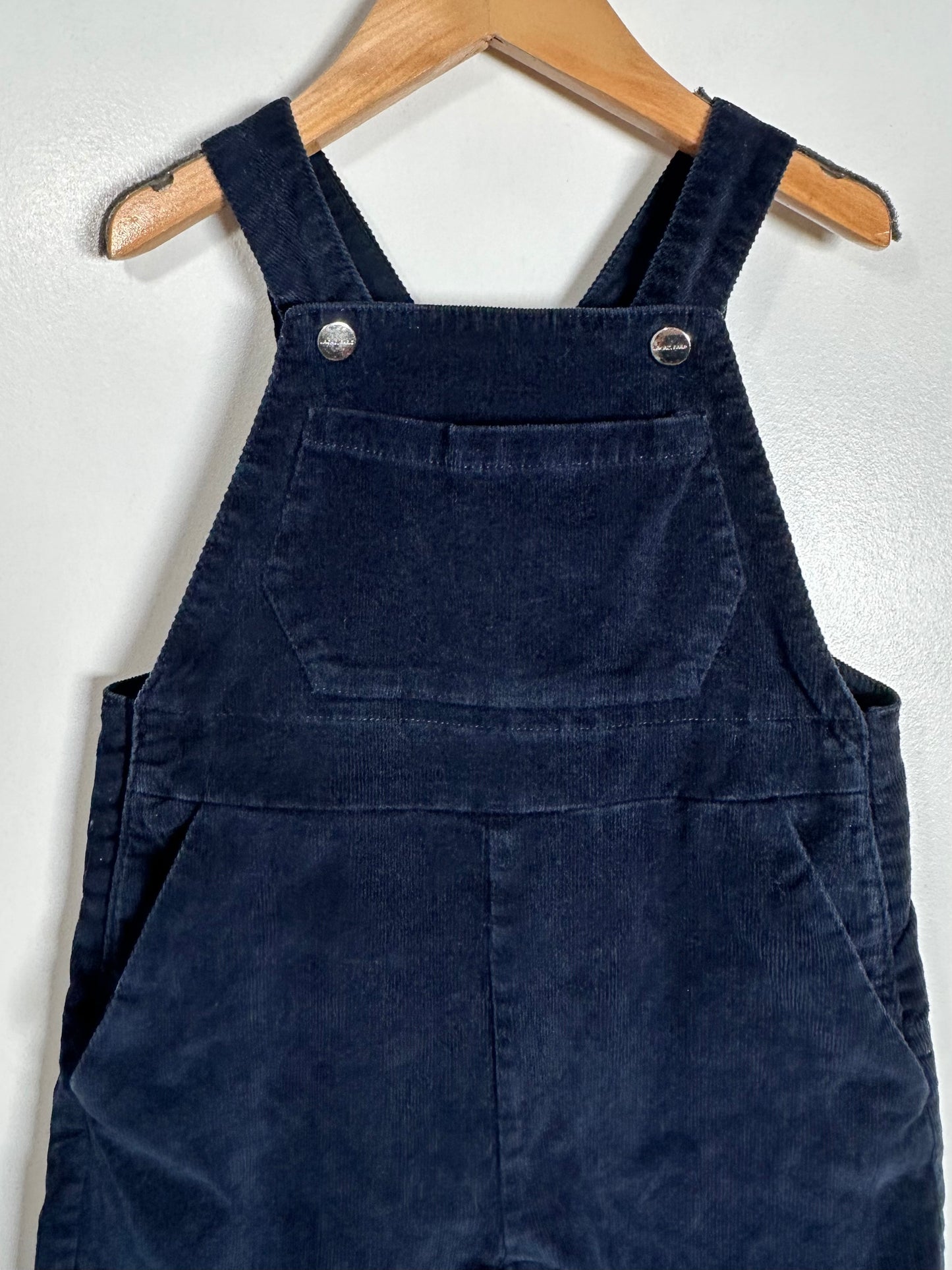 A pair of dark navy blue corduroy bib overalls hanging on a wooden hanger against a white wall.