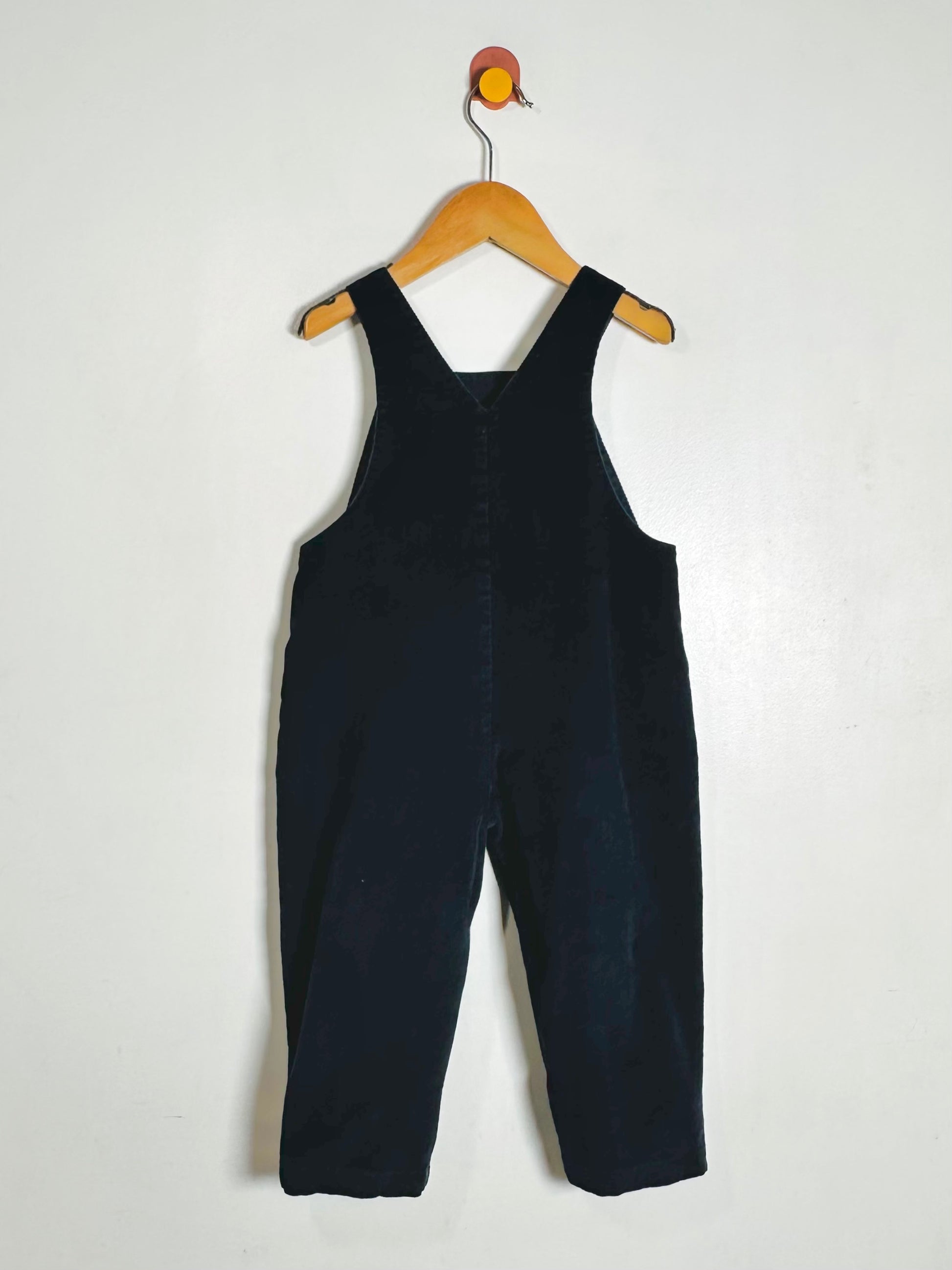 A dark navy corduroy baby overall jumpsuit hanging on a wooden hanger against a plain off-white wall.