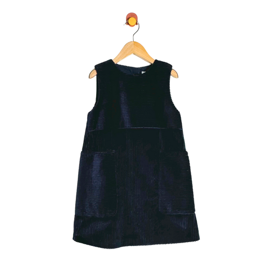 A sleeveless dark navy corduroy dress with front patch pockets, hanging on a wooden hanger against a plain white background.