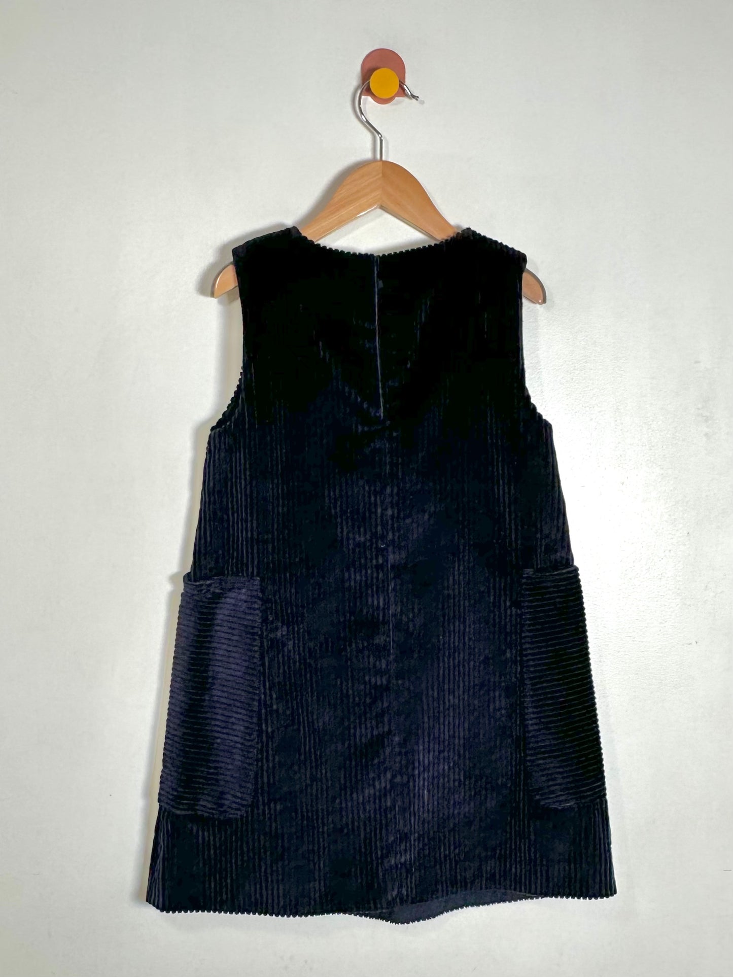 A sleeveless dark navy corduroy dress or pinafore hanging on a wooden hanger against a plain off-white wall.