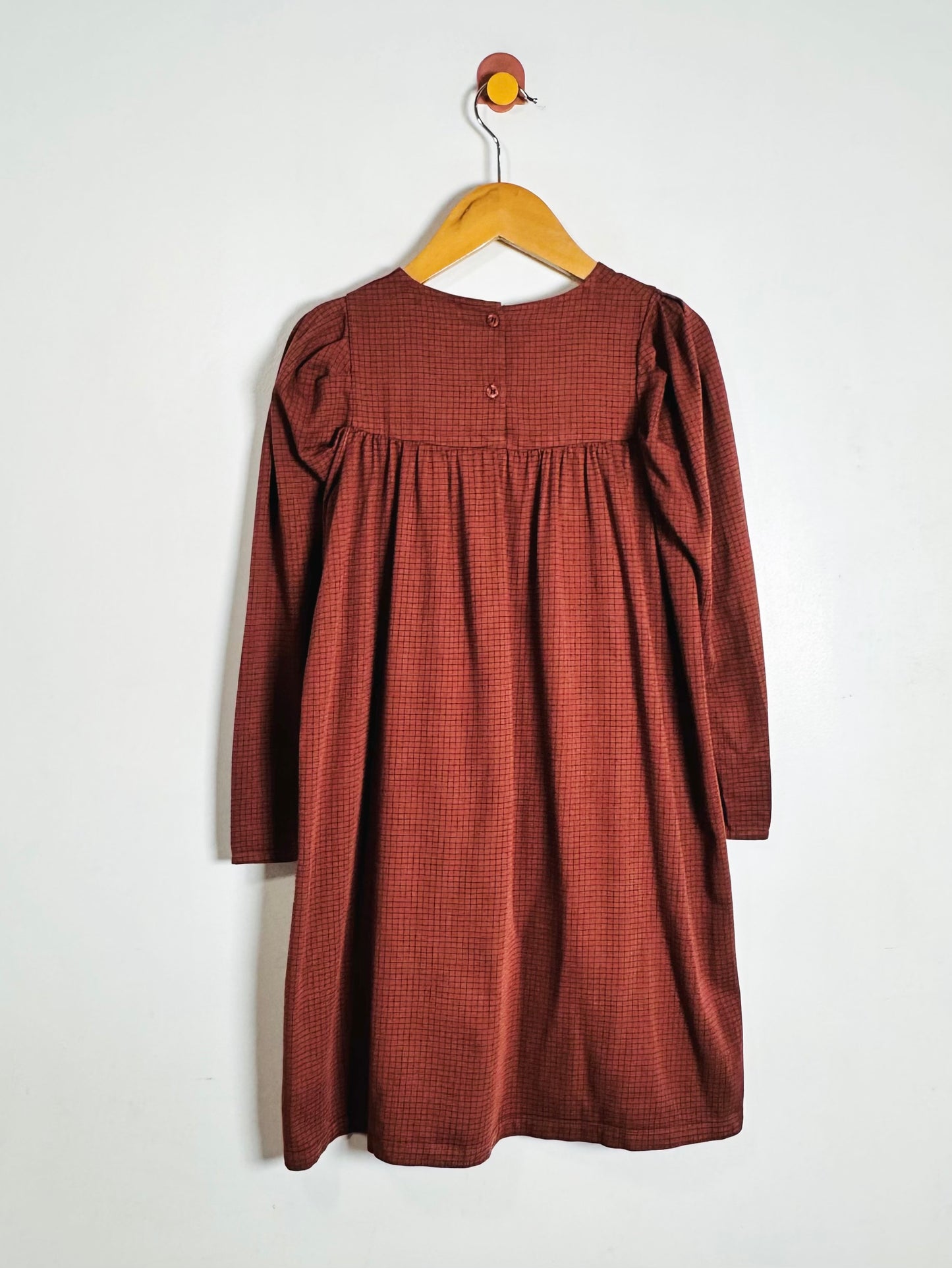 A rust-colored long-sleeve baby dress with a small checkered pattern, hanging on a wooden hanger against a plain white wall.