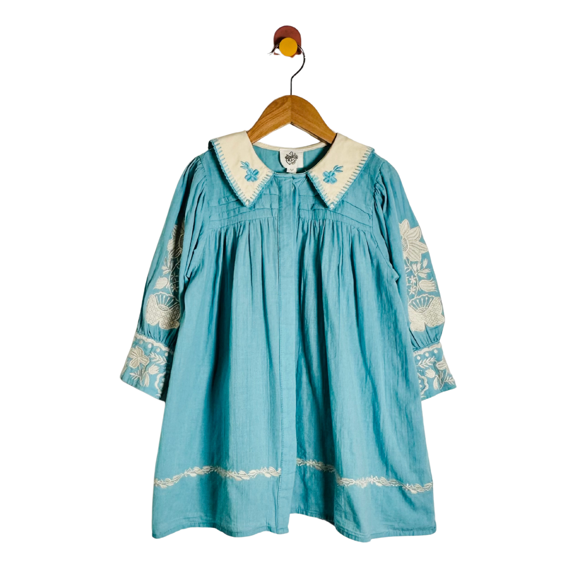 A light blue denim-style dress with cream embroidery on the collar, cuffs, and hem, hanging on a wooden hanger against a white background.
