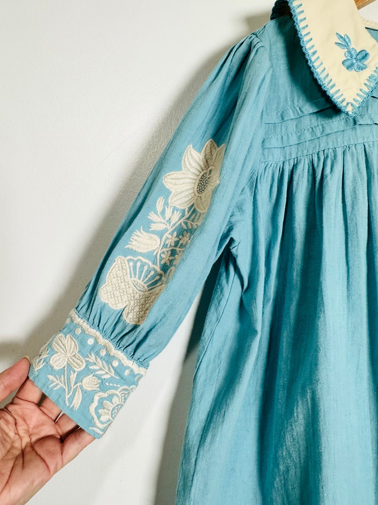 A close-up of a teal blue long-sleeve dress with cream floral embroidery on the sleeve and cuff, featuring a cream collar with blue embroidery, held by a hand against a plain white wall.