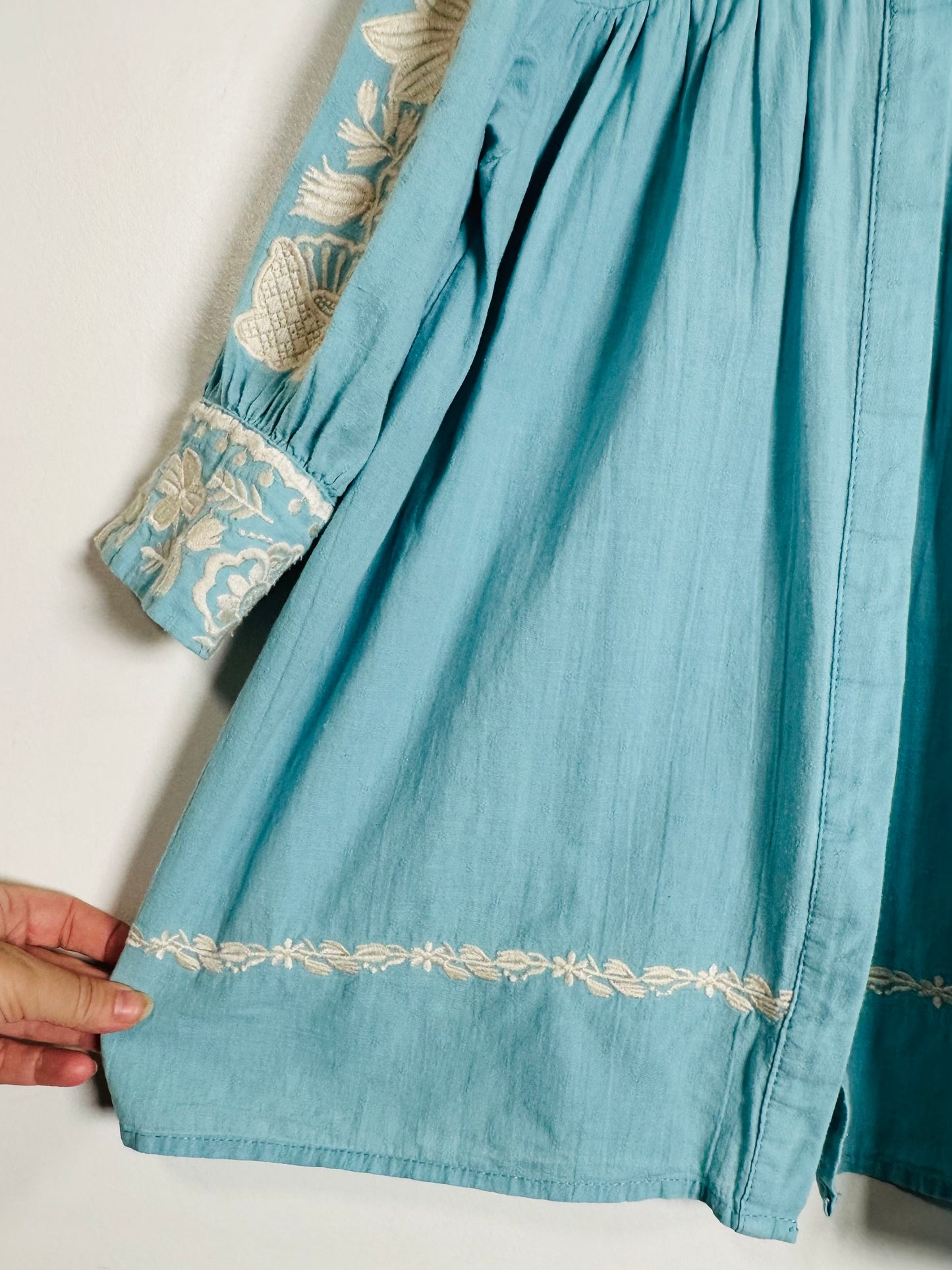 A close-up of a light blue cotton dress or tunic with cream-colored floral embroidery on the sleeve cuff and hem, held by a hand against a plain white background.