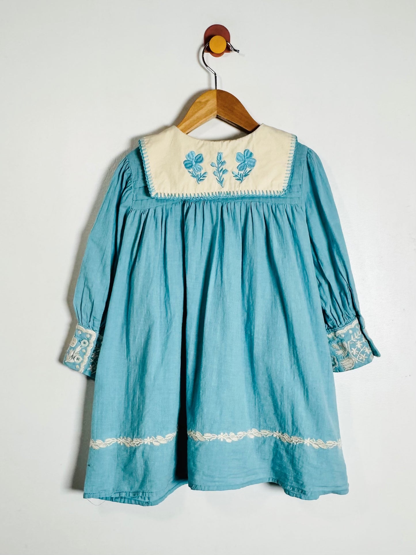A light blue long-sleeve baby dress with a cream-colored embroidered collar and cuffs, hanging on a wooden hanger against a plain white wall.
