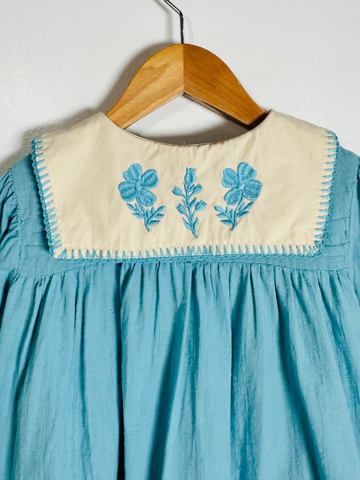 A light blue cotton dress hanging on a wooden hanger against a white wall, featuring a cream-colored collar with blue embroidered flowers and green leaves.