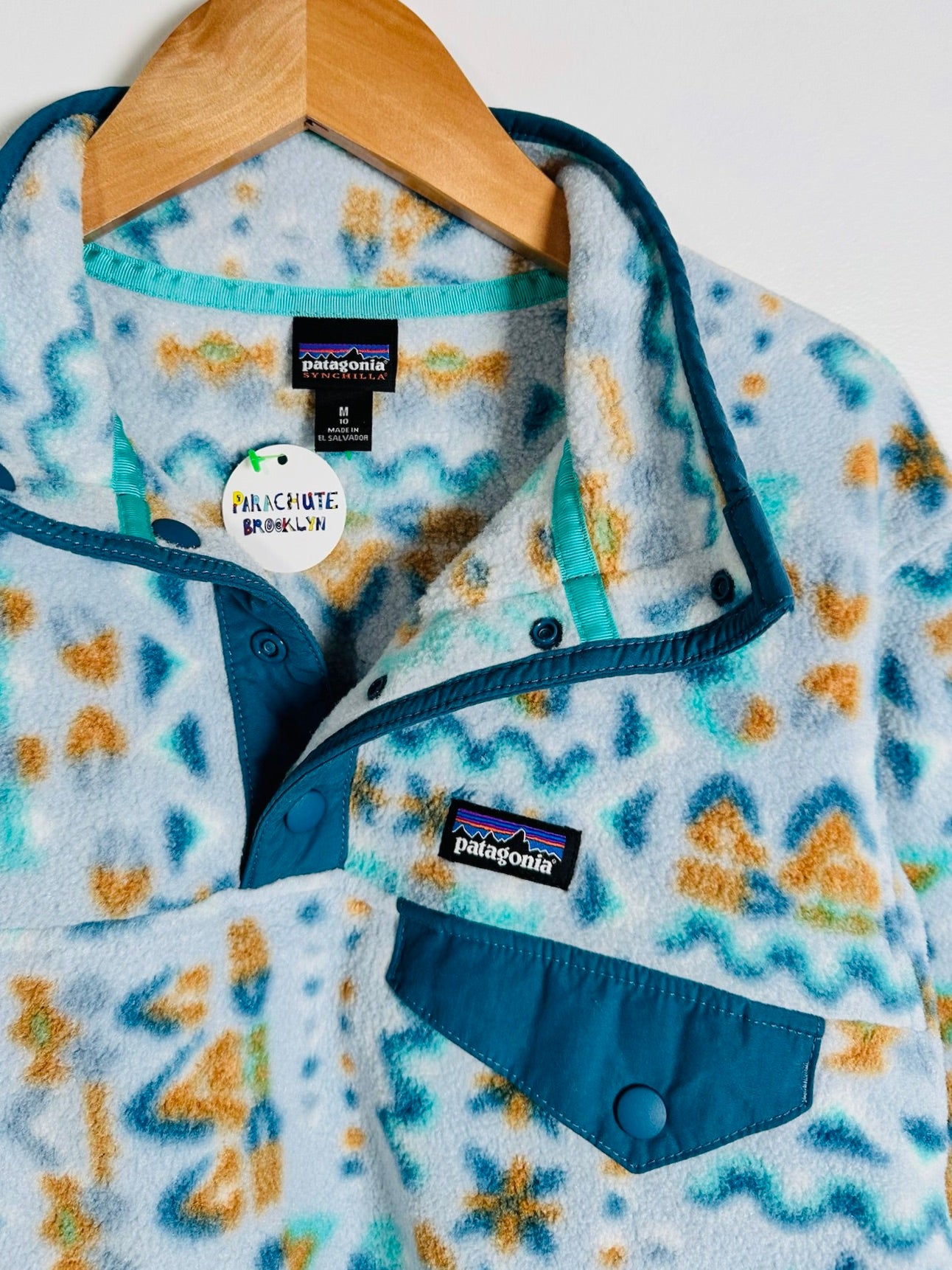 A close-up of a Patagonia fleece pullover shirt on a wooden hanger, featuring a light blue and white tie-dye pattern with teal and orange accents, a teal collar, and a front chest pocket.