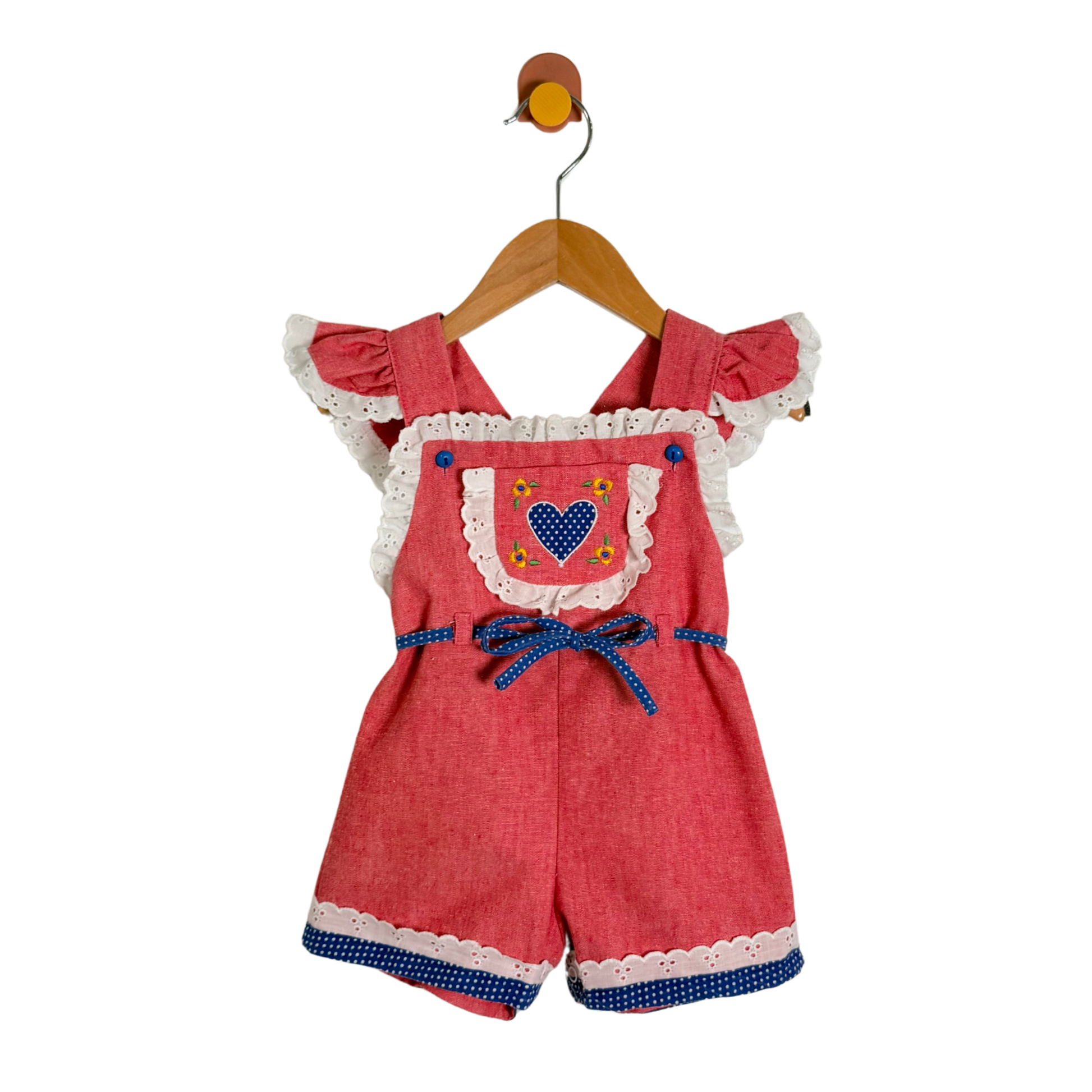 A baby girl's short romper in a textured coral-red fabric, featuring white lace trim, ruffled sleeves, a blue polka-dot bow at the waist, and a chest pocket with a blue heart design. The romper is hanging on a wooden hanger against a plain white background.