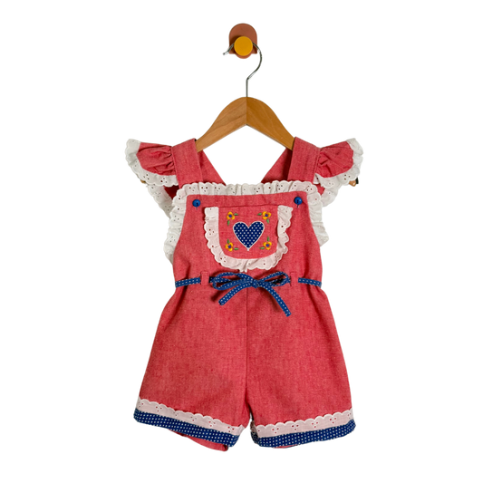 A baby girl's short romper in a textured coral-red fabric, featuring white lace trim, ruffled sleeves, a blue polka-dot bow at the waist, and a chest pocket with a blue heart design. The romper is hanging on a wooden hanger against a plain white background.